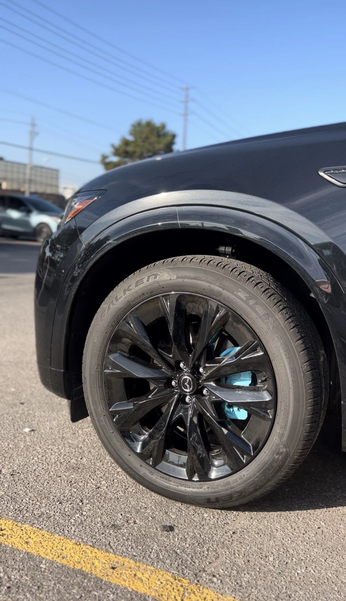 Close-up of a black Mazda car wheel with a Falken tire and blue brake caliper, parked in a parking lot with other vehicles and a clear blue sky.