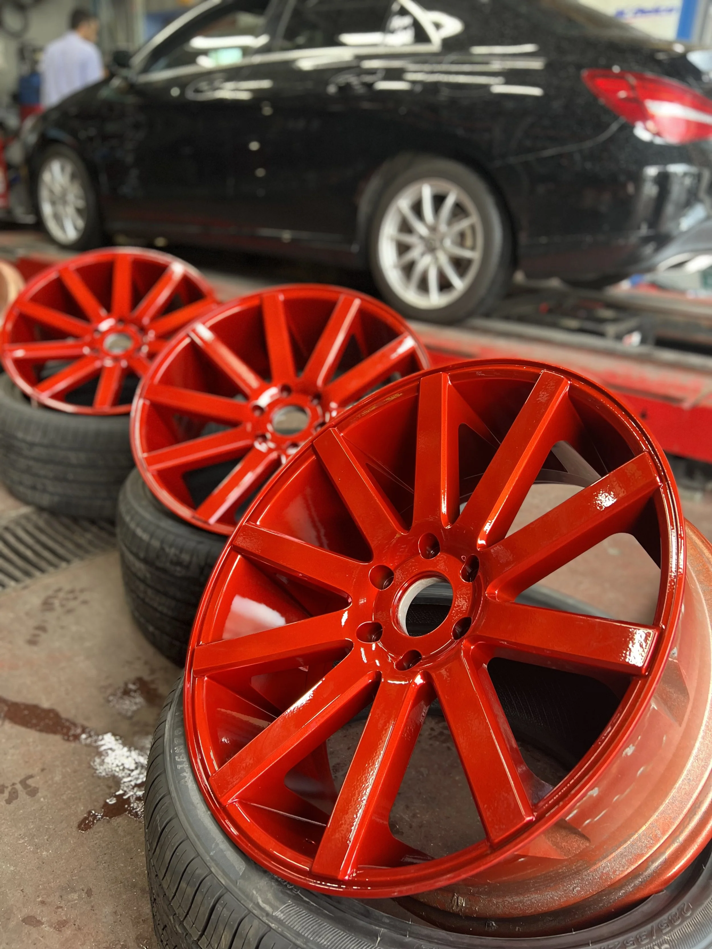 Three red alloy car wheels stacked on top of tires with a black car in the background in a garage or auto shop.