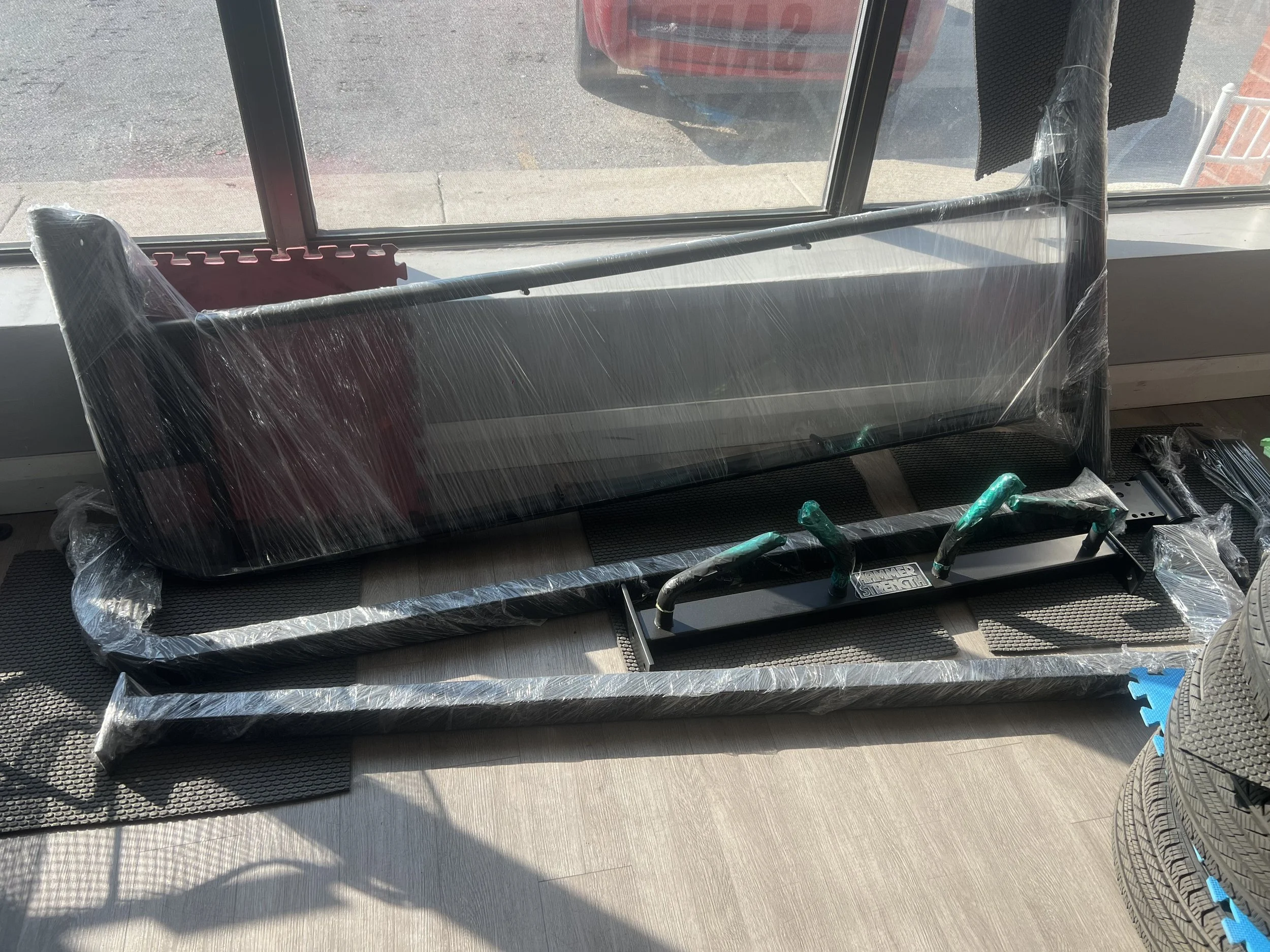 Various car parts, including a large black rear spoiler, a bumper, and some tools, are wrapped in plastic and placed on the floor near a large window at an auto shop or garage.