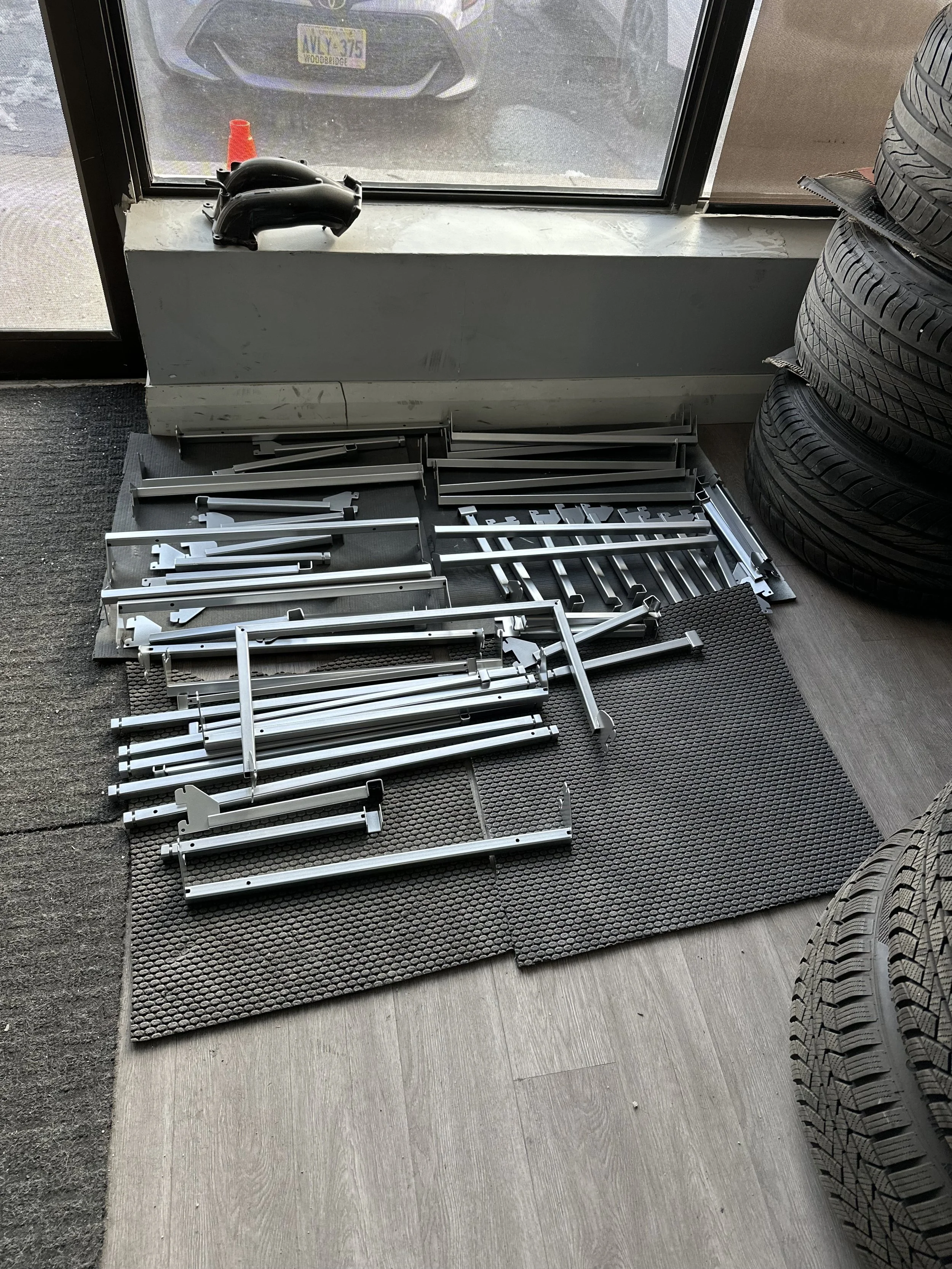 Disassembled parts of a car interior, including metal rails and brackets, on black rubber mats near a window in an automotive shop.