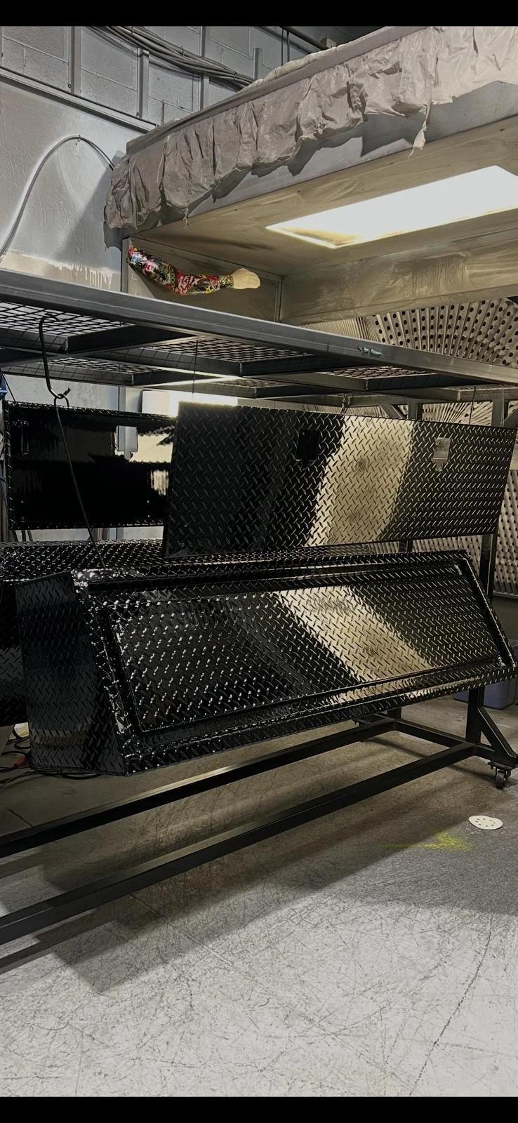 A black metal grill with a diamond pattern sits on a wheeled stand in a workshop or industrial space, with various metal racks and a sheet of plywood behind it.