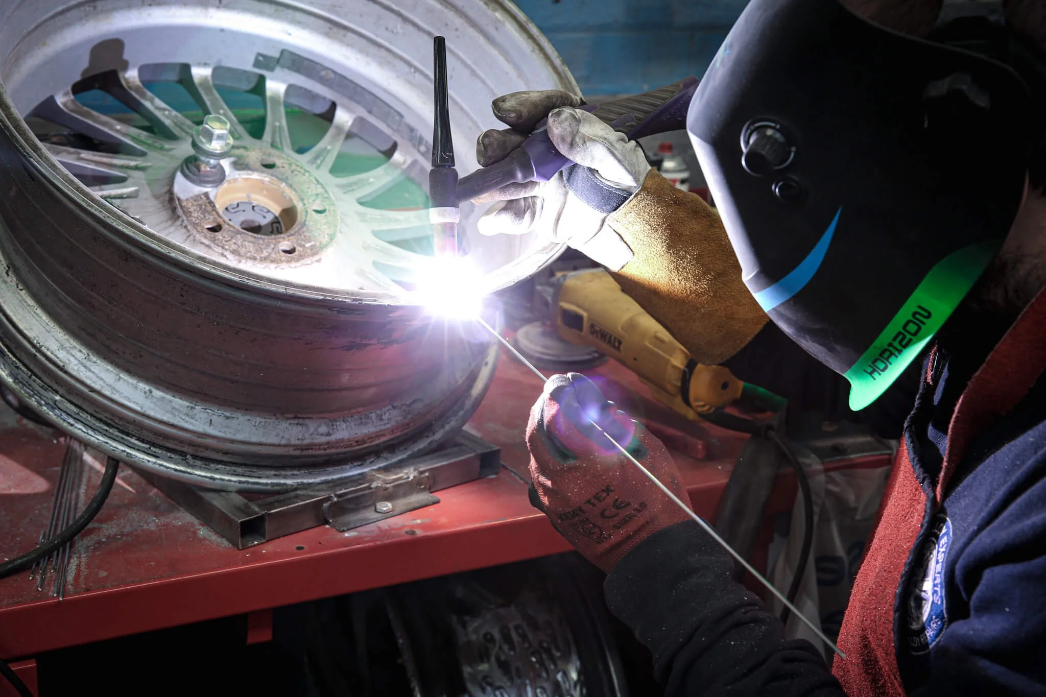 A person working on a car wheel in a garage or workshop, with tools and equipment visible in the background.