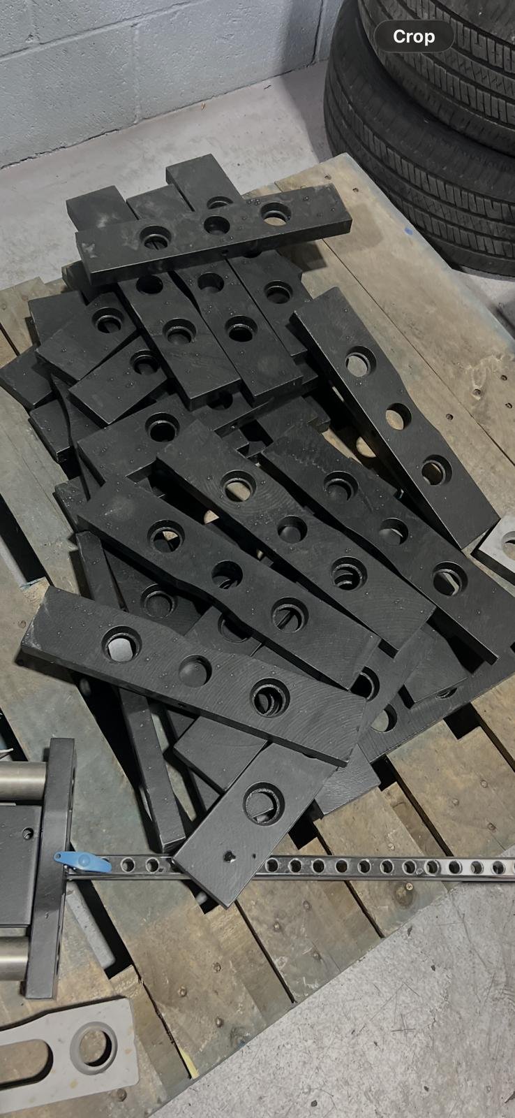 Stack of black metal plates with circular holes on a wooden pallet, next to a set of car tires.