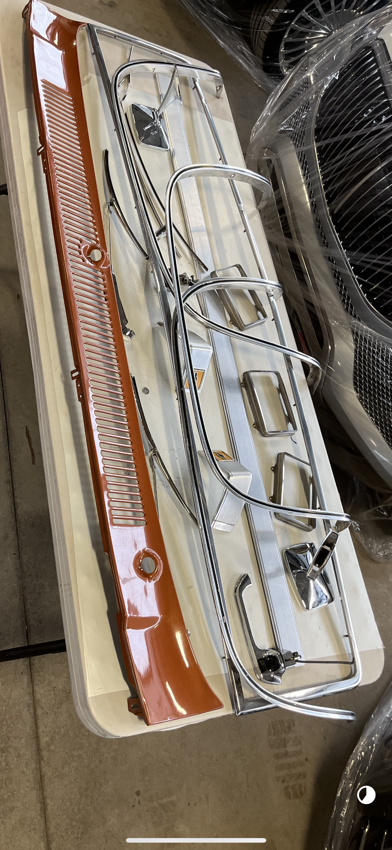 A collection of metal and plastic parts, possibly from a vehicle, laid out on a white surface. The parts include metal bars, handles, brackets, and a reddish-brown plastic component. In the background, there are stacked car wheels wrapped in plastic.