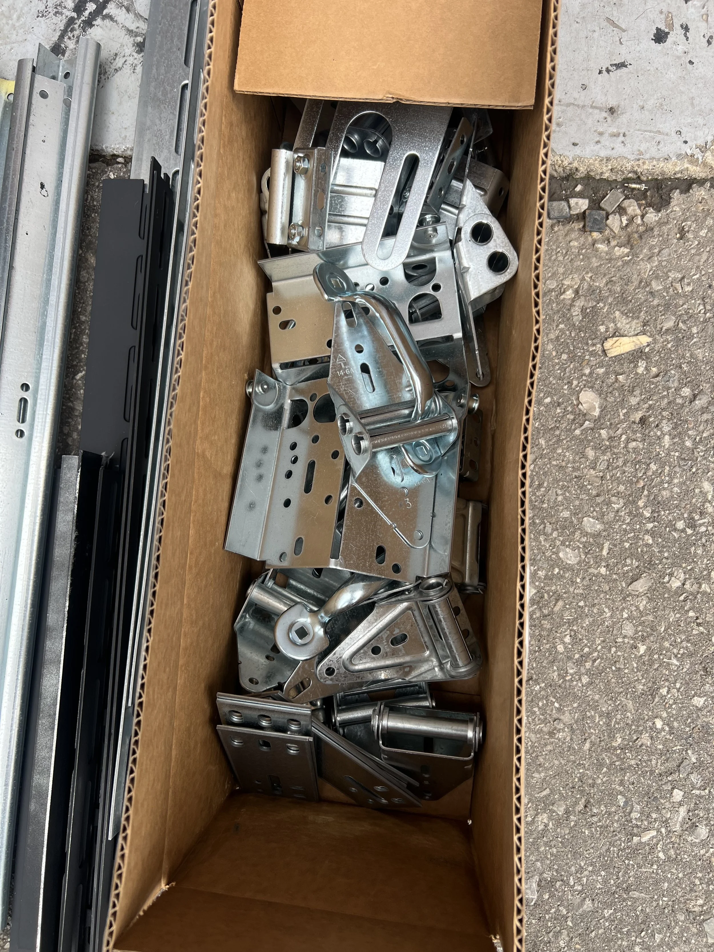 A cardboard box filled with metal construction brackets and hardware parts, placed outdoors on a gravel surface.