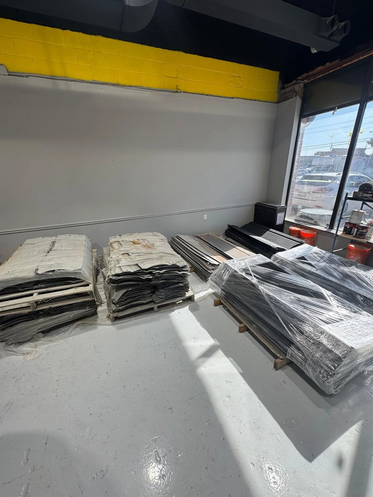 Piles of large, flattened cardboard and paper stacked and wrapped in plastic in a room with large window sunlight, gray walls, and a yellow strip near the ceiling.