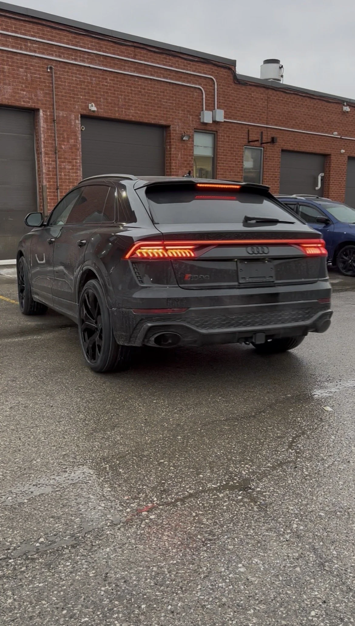 Black Audi RS Q5 parked in front of a brick building with gray garage doors and a blue car parked nearby.