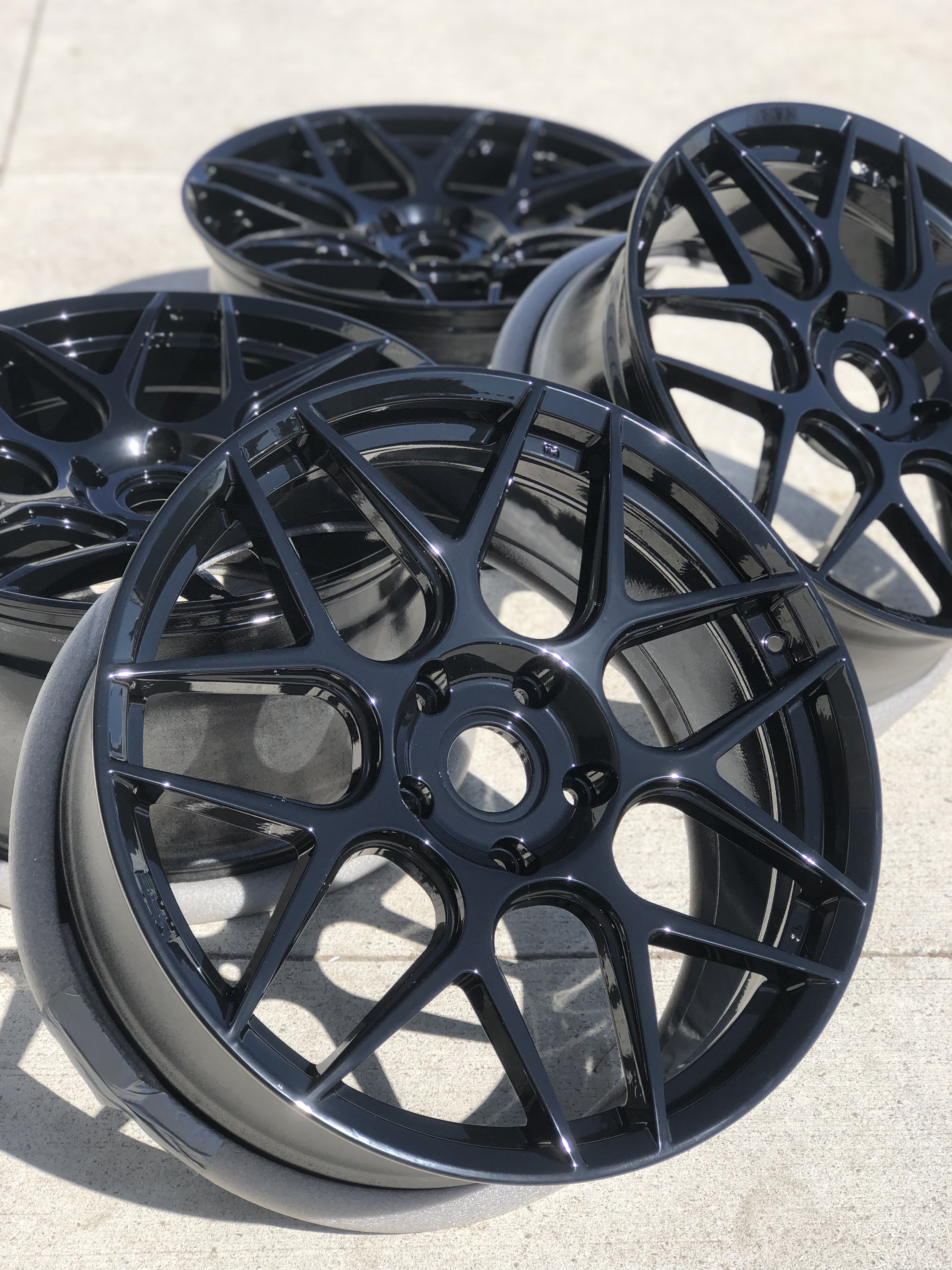 Four black, multi-spoke car wheels positioned on a concrete surface with sunlight reflecting off their glossy finish.