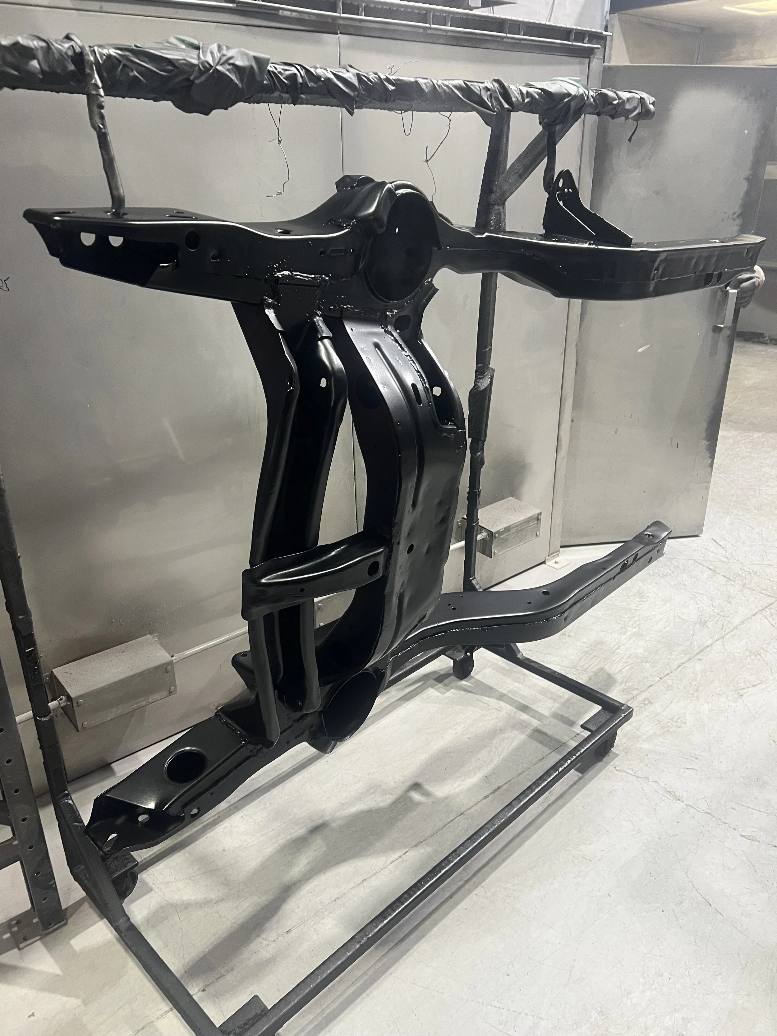 A black painted metallic frame of a vehicle, possibly part of a chassis or structural component, mounted on a metal frame stand in a workshop or manufacturing environment.