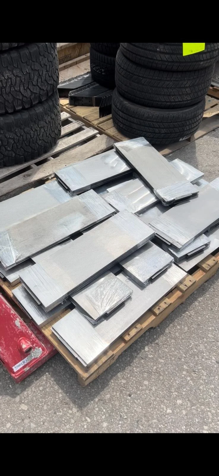 Stacked metal tiles on a wooden pallet with car tires in the background.
