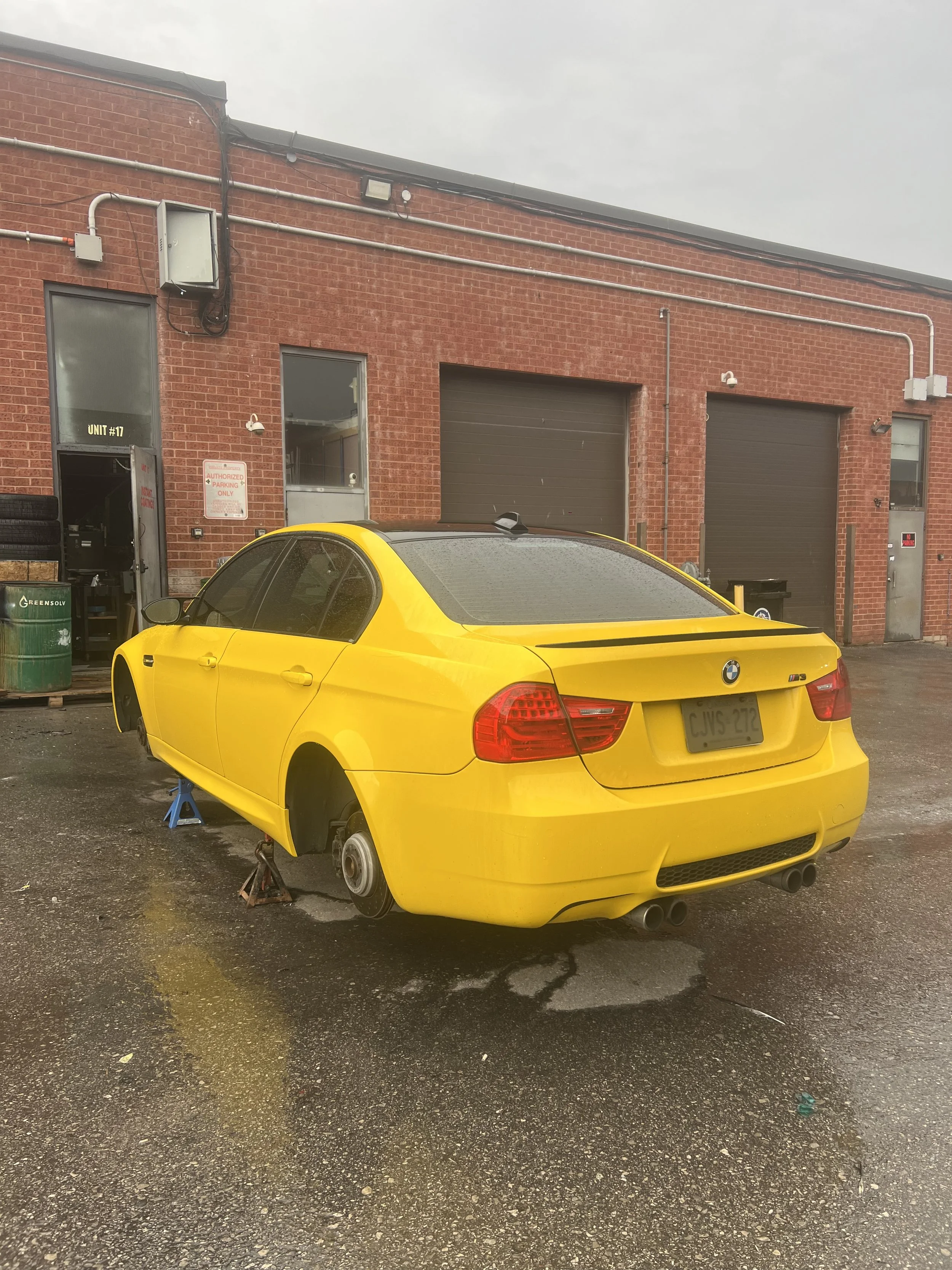 Yellow BMW M3 car on jack stands in an industrial parking lot with brick building in background.
