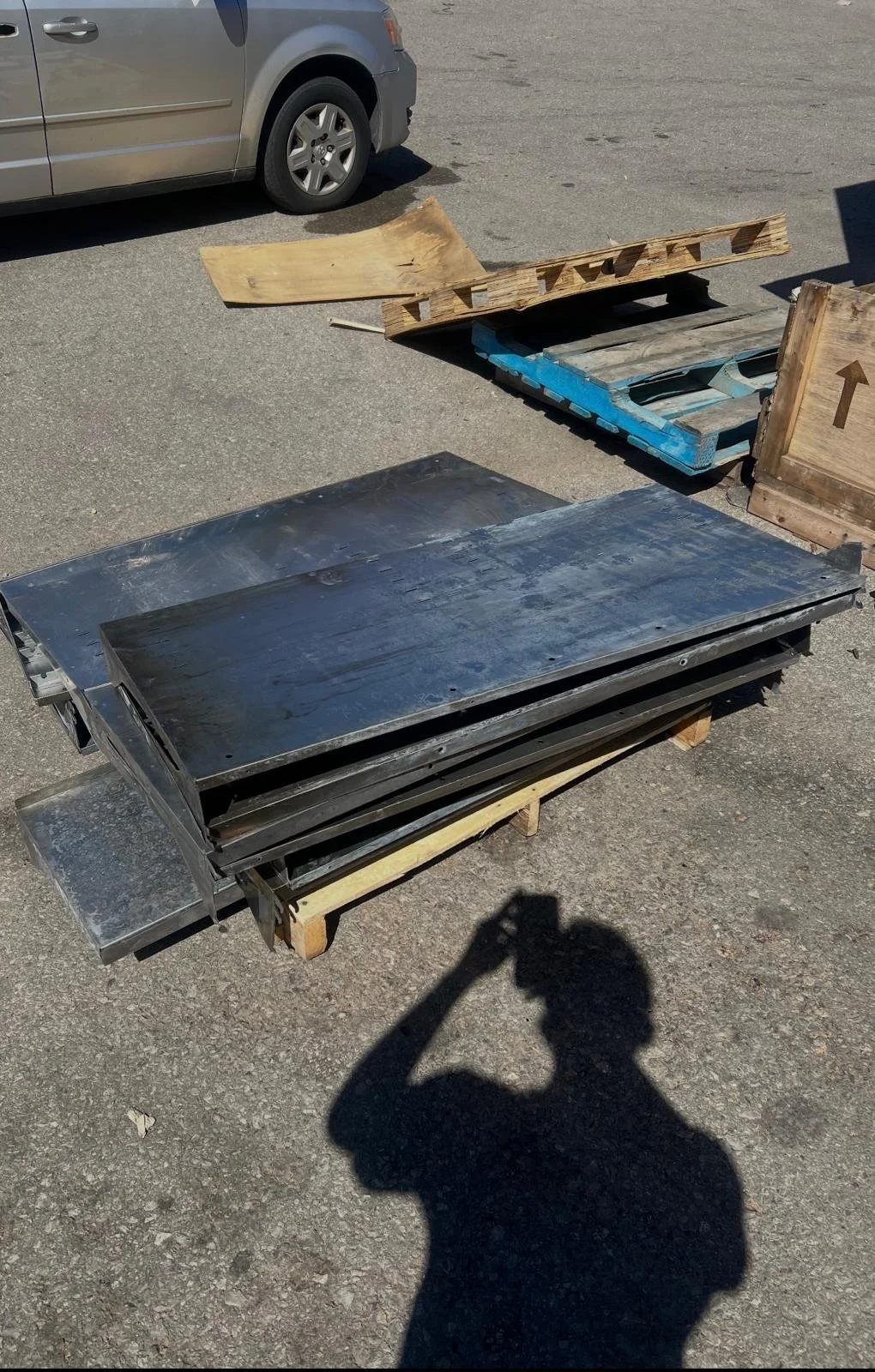 Various industrial materials and pallets on an outdoor surface, with a car in the background and the photographer's shadow in the foreground.