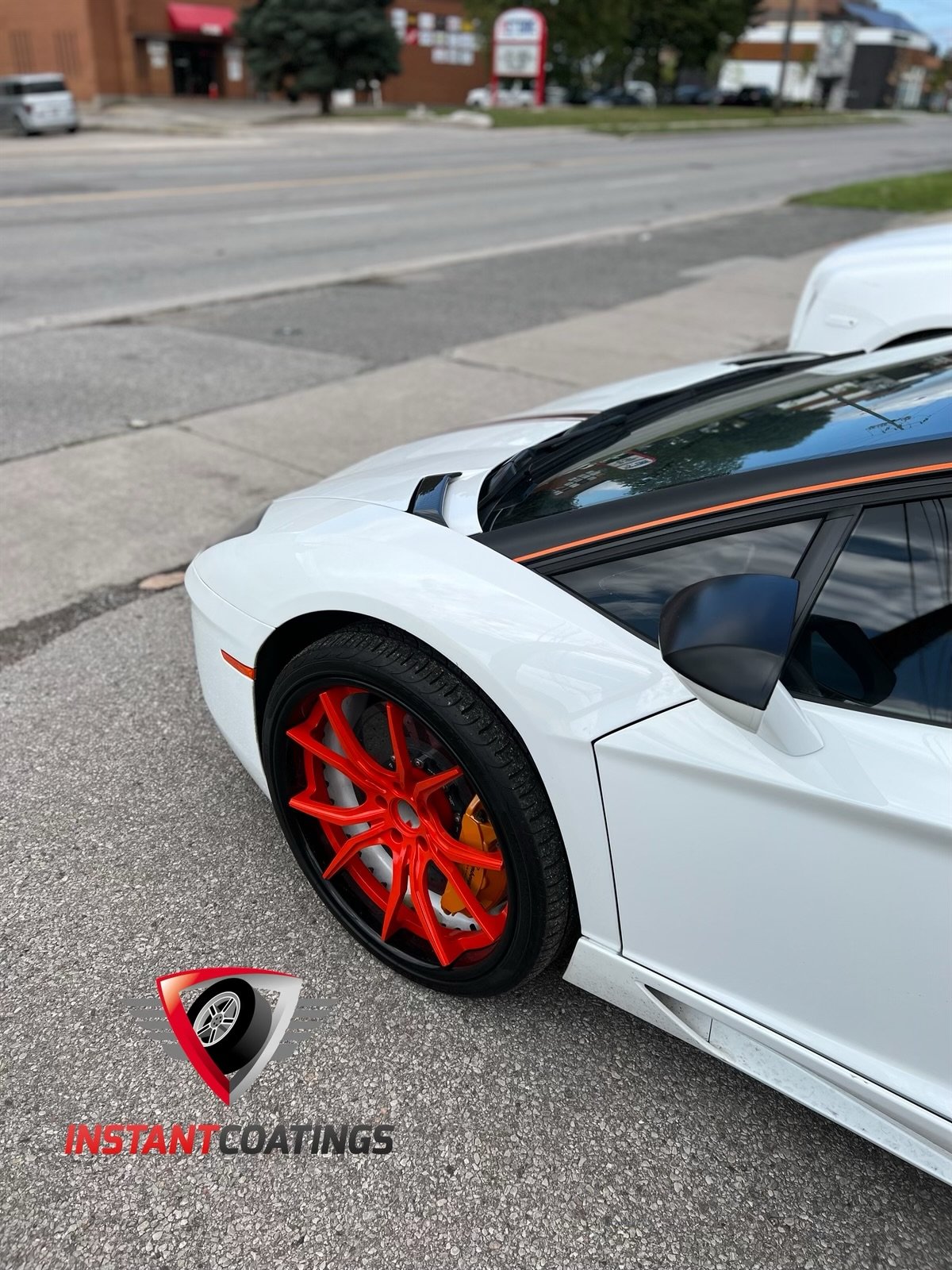 Close-up of a white sports car with striking red wheels parked on a street, with a business and trees in the background.