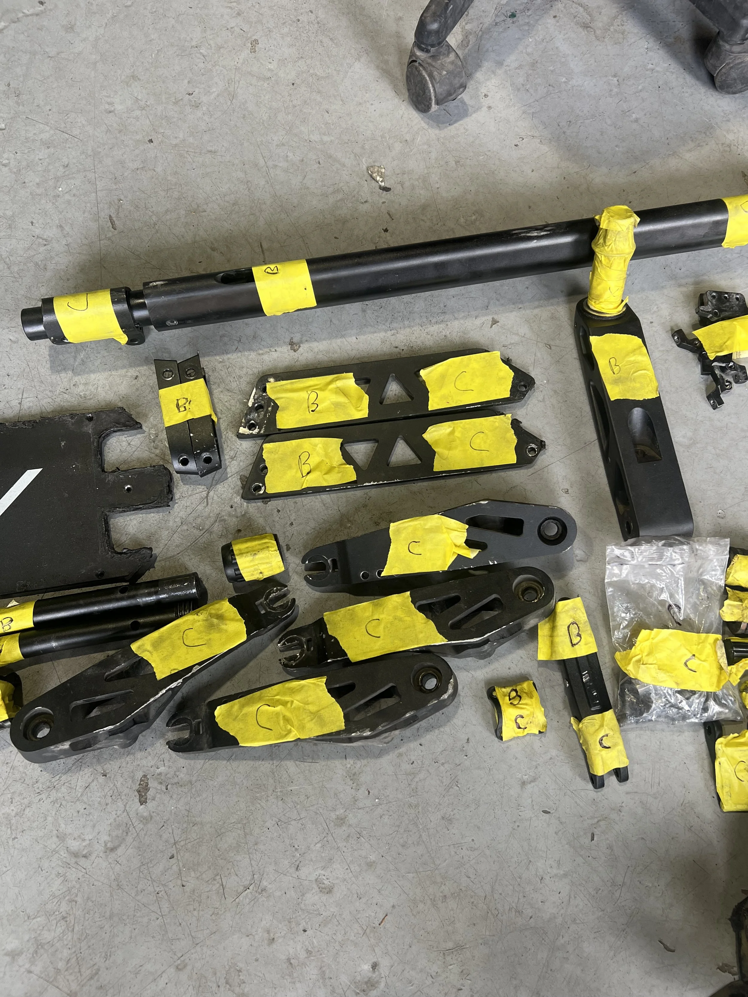 Disassembled motorcycle parts laid out on a concrete floor, each labeled with yellow tape marked with letters.