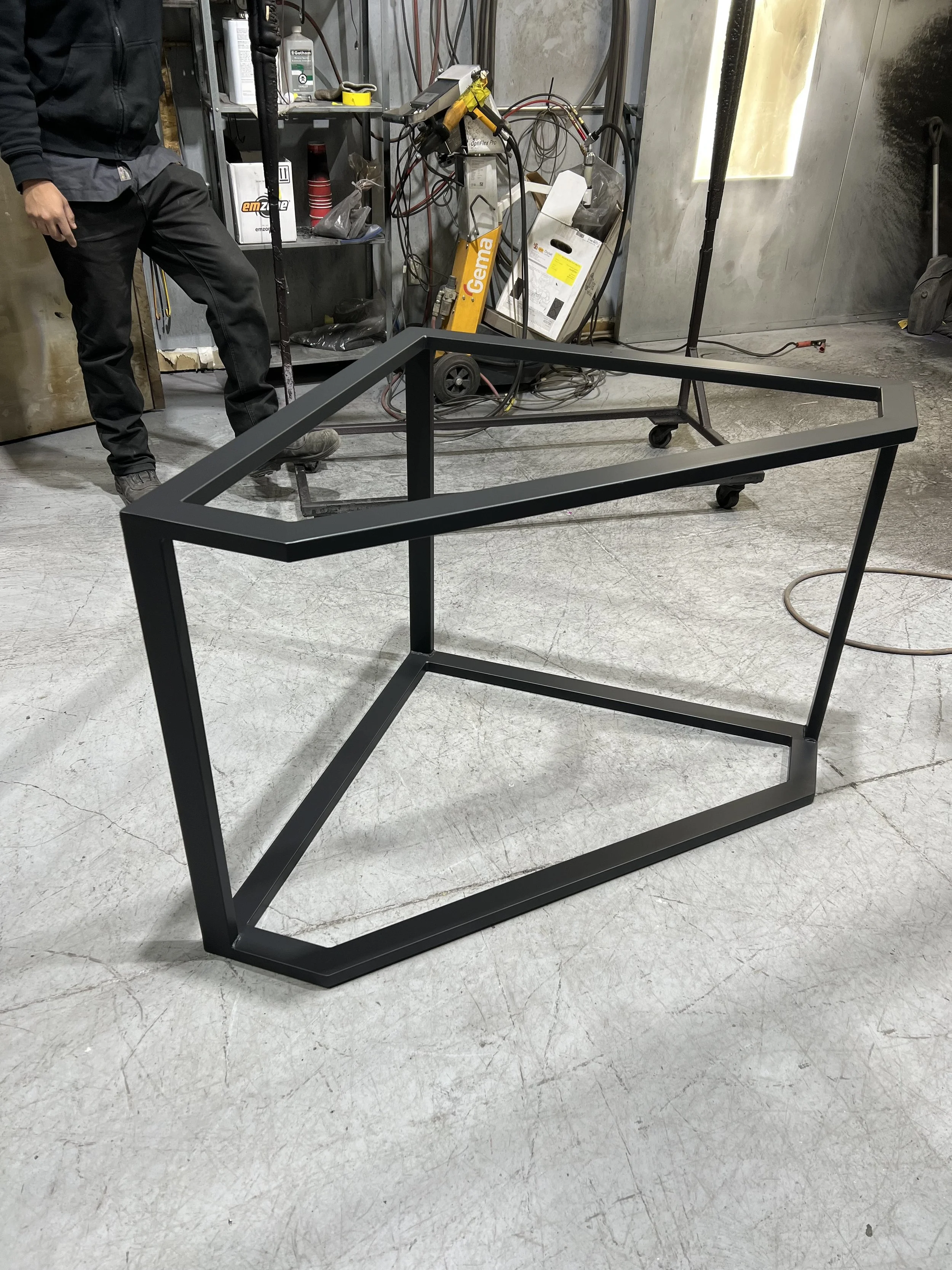 A black metal frame for a piece of furniture in a workshop or garage. In the background, there are tools, equipment, and a person partially visible.