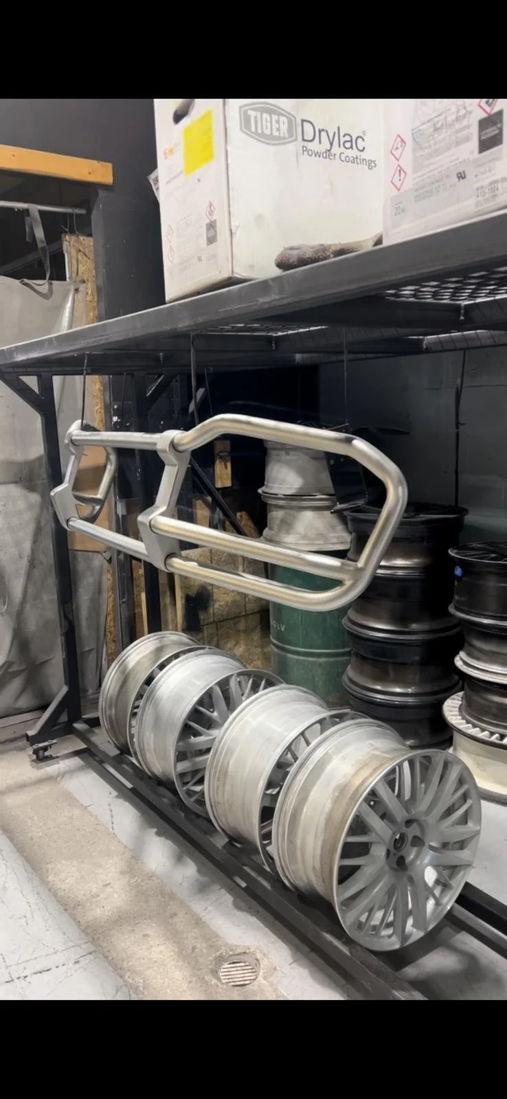 Set of car rims and a tire lock spacer on a black metal rack, with boxes and metal parts in the background.