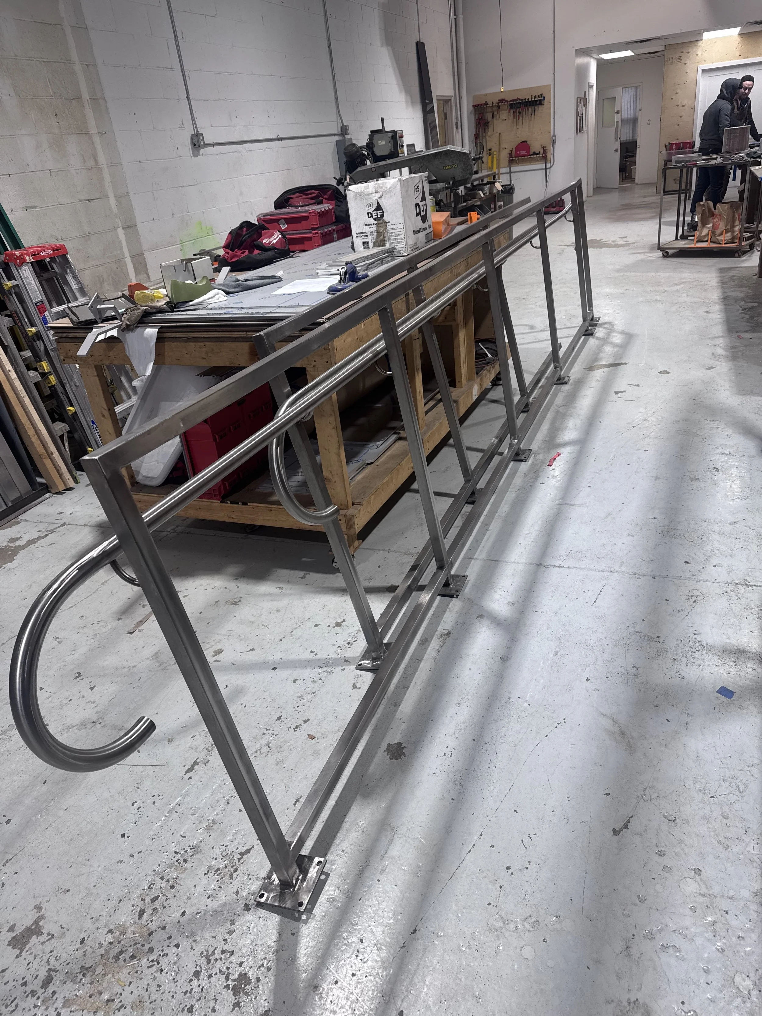 A metal wheelchair ramp leans against a workbench in a workshop with tools, boxes, and two people working in the background.