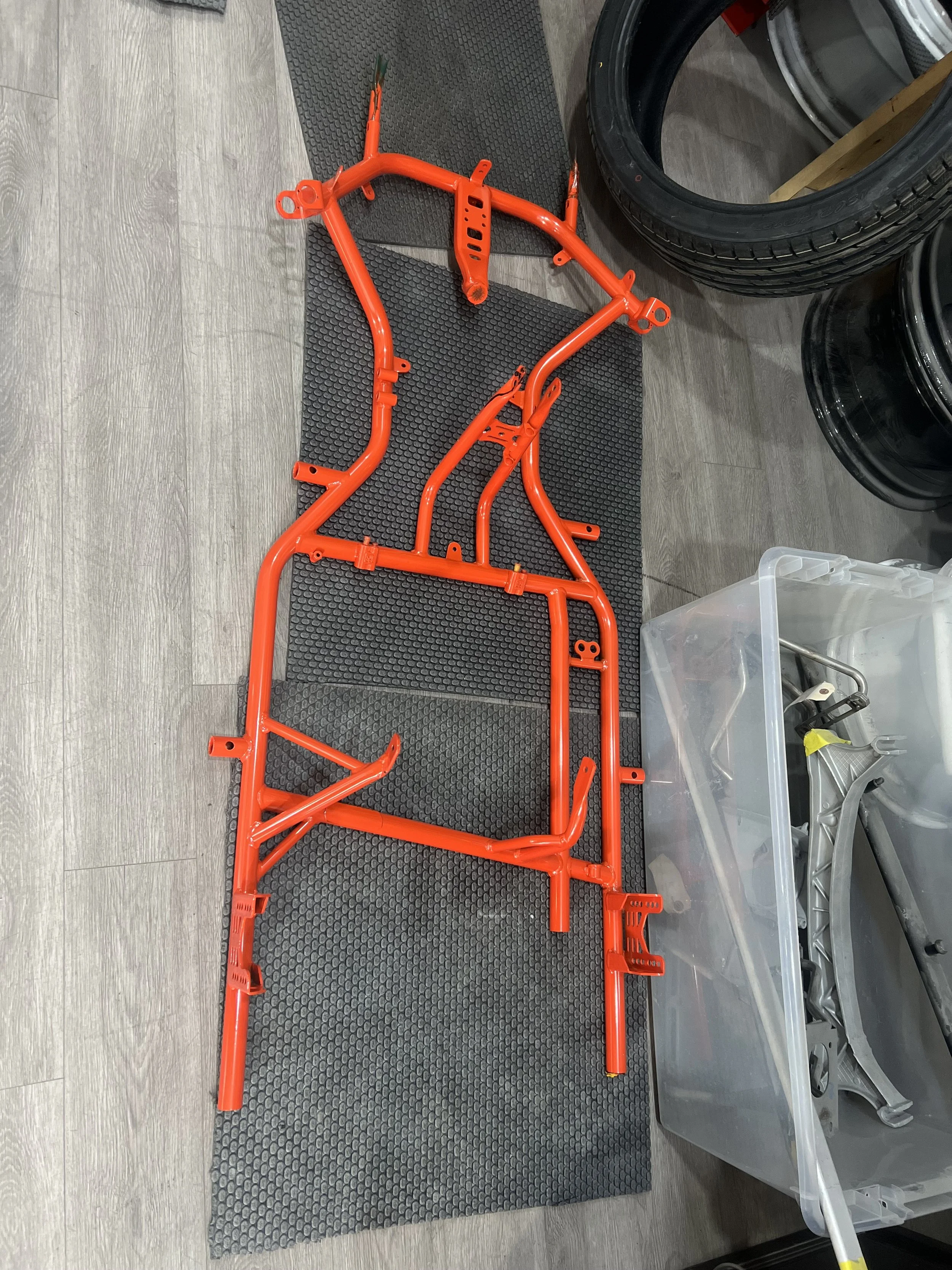 Red-painted metal motorcycle frame lying on black rubber mats, with tires and other motorcycle parts and tools nearby.