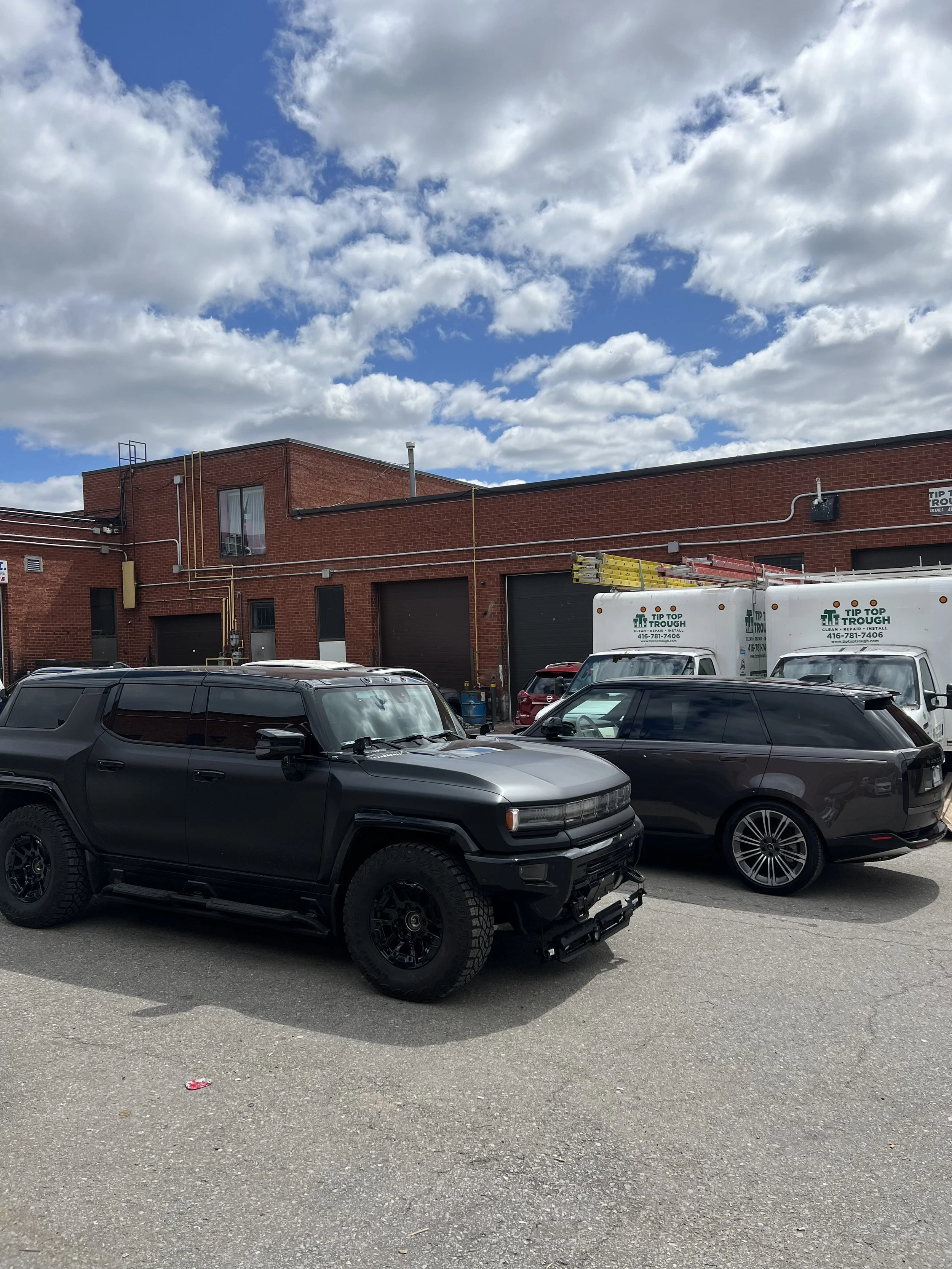 Parking lot with black SUV, gray station wagon, and commercial trucks in front of a red-brick industrial building under partly cloudy sky.