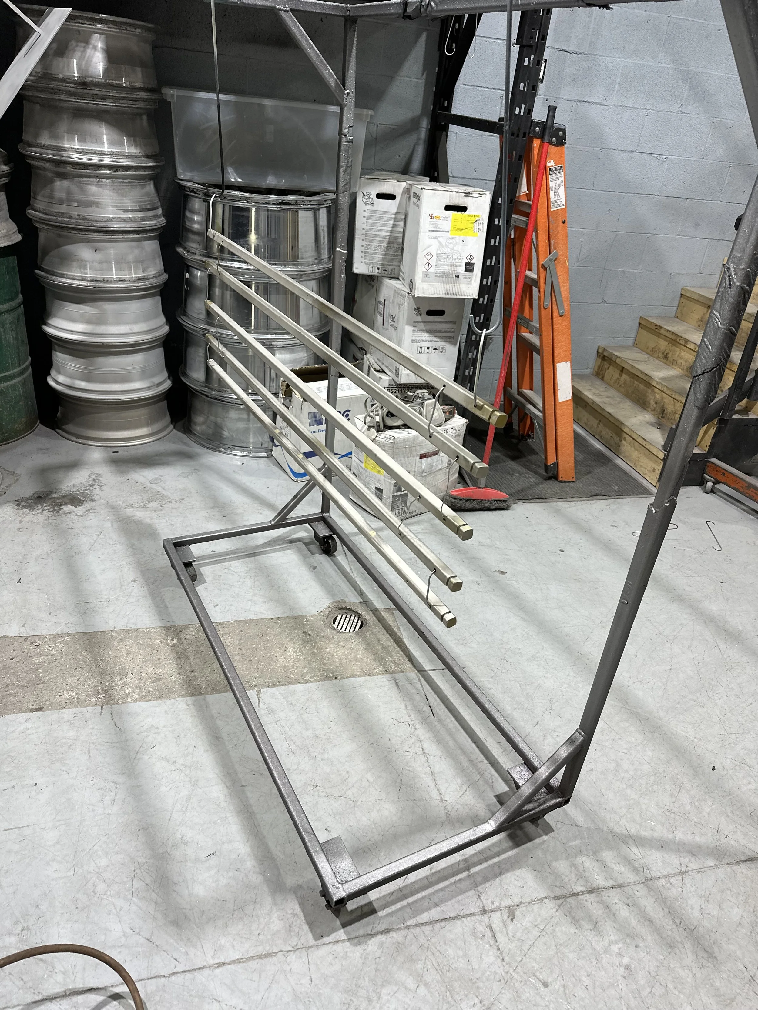 Empty metal laundry cart with three horizontal bars and wheels, on a garage concrete floor, surrounded by stacked metal rims, boxes, a ladder, and stairs.