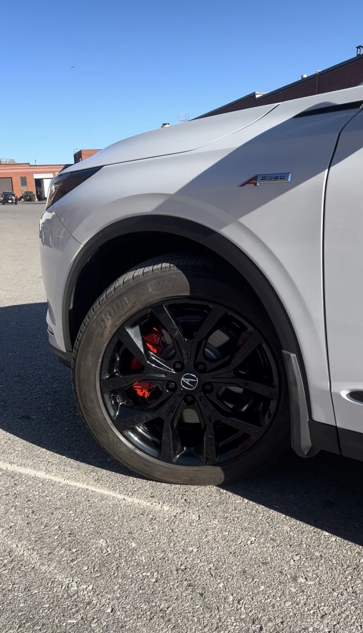 Close-up of the front part of a silver Honda SUV, including the wheel, tire, brake caliper, and part of the front bumper, with a clear blue sky in the background.