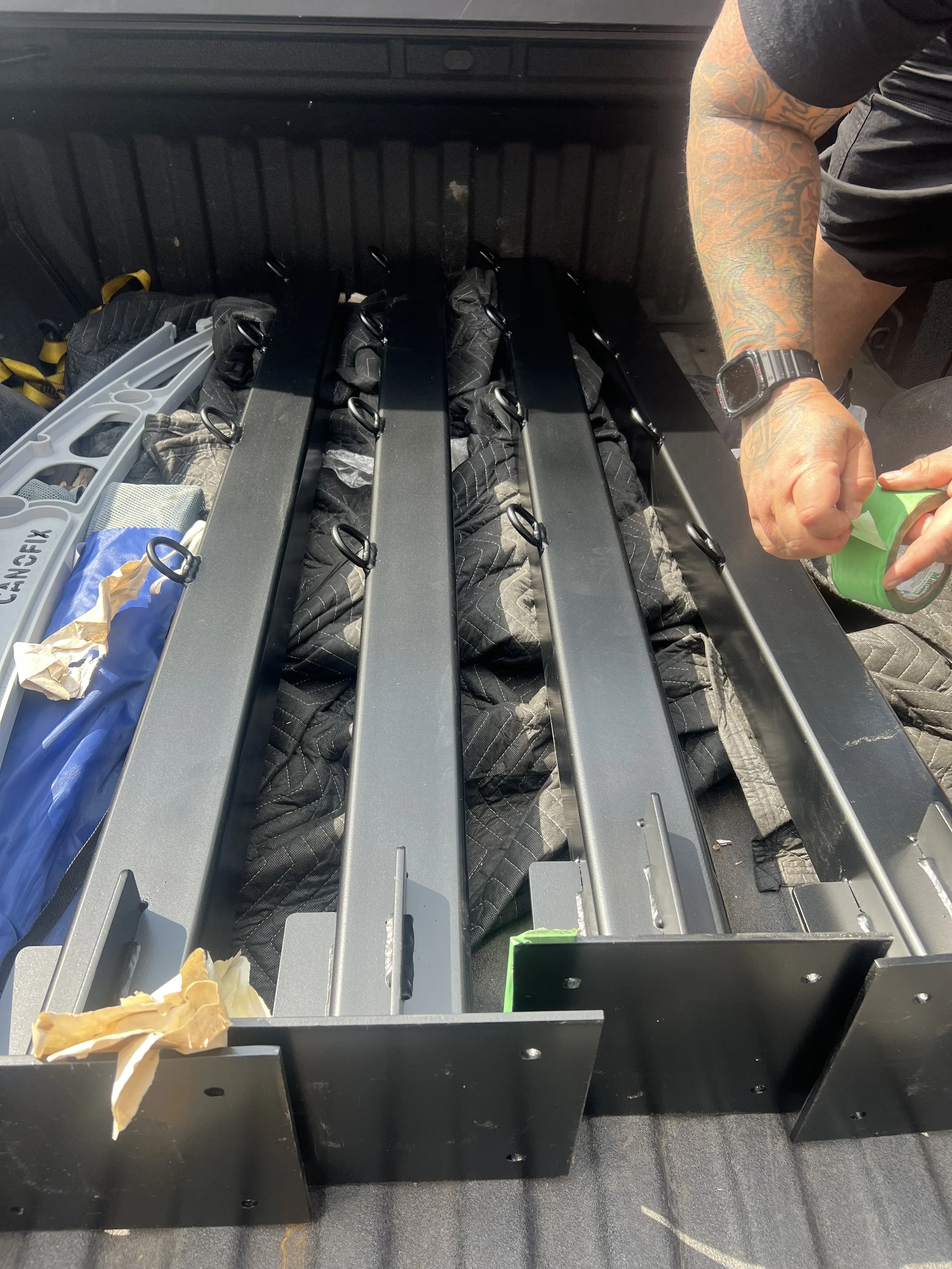 Person with tattooed arms is working with tools and green painter's tape in the bed of a pickup truck, which contains some black quilted fabric, cardboard, and various equipment.