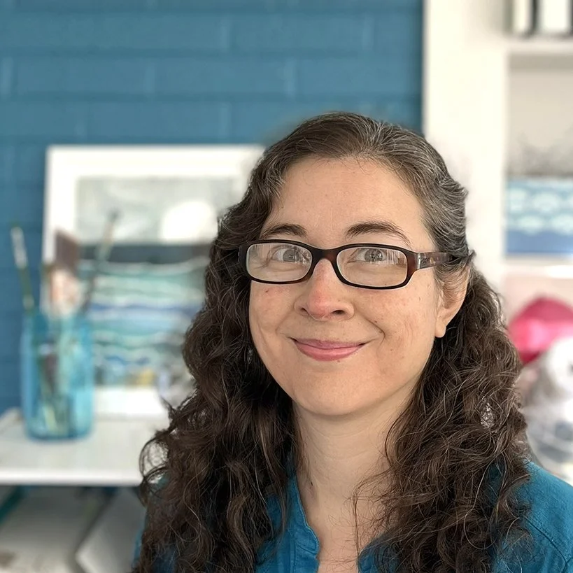 A woman with long curly hair, glasses, and a blue shirt smiling at the camera in an office or studio setting.