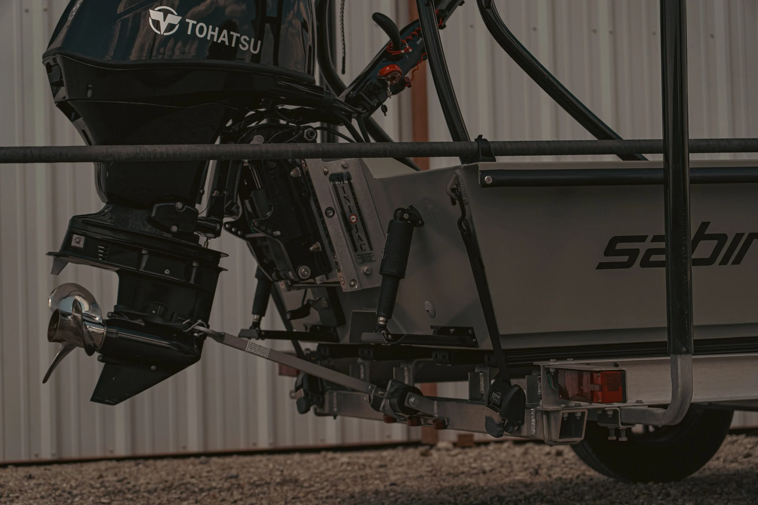 A boat on a trailer being pulled by a vehicle, with an outboard motor attached at the stern. The boat has the word 'Sabre' on its side and is parked on a gravel surface next to a large, light-colored industrial building.