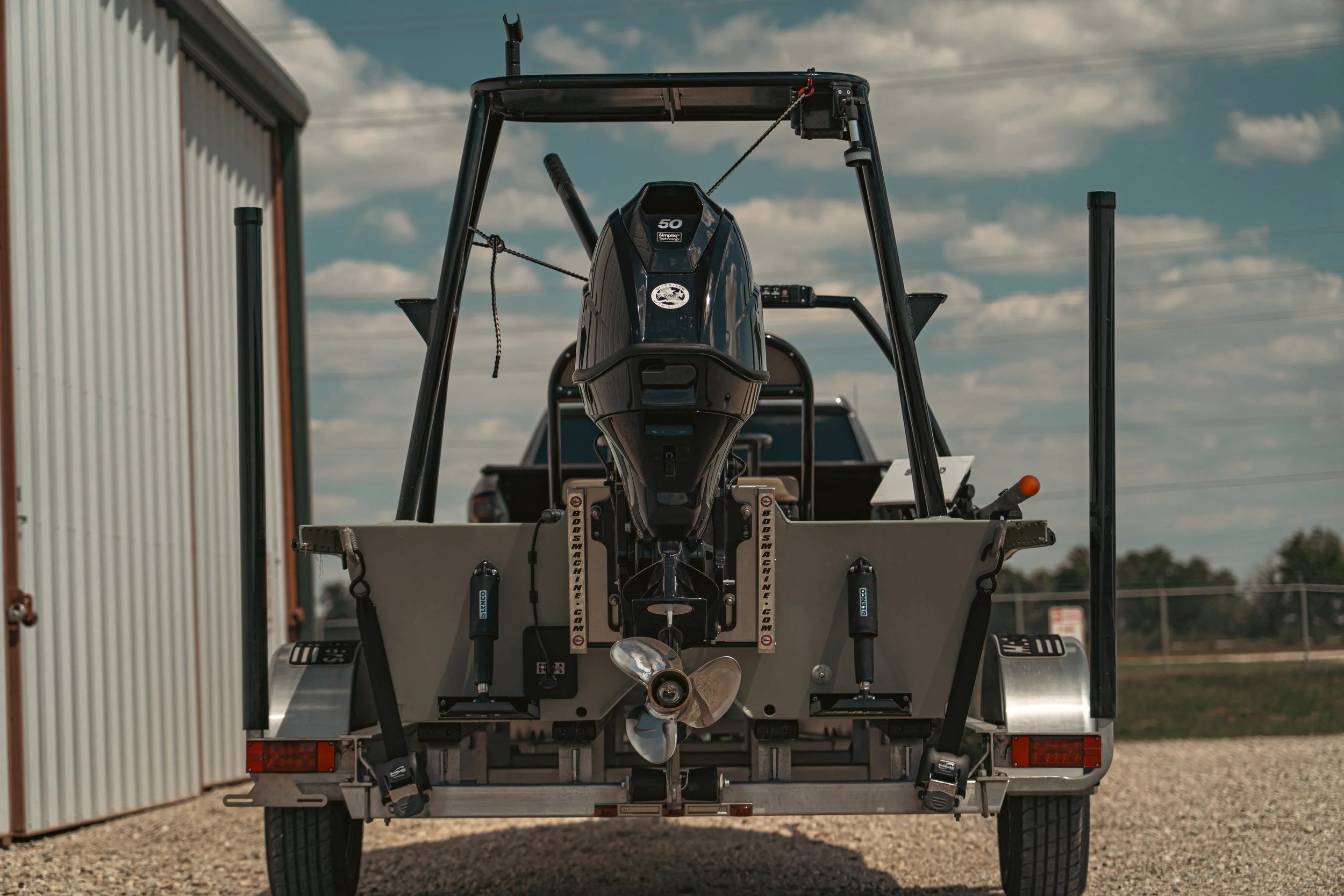 A boat with a powerful outboard motor on a trailer parked outdoors near a metal building, with a partly cloudy sky in the background.
