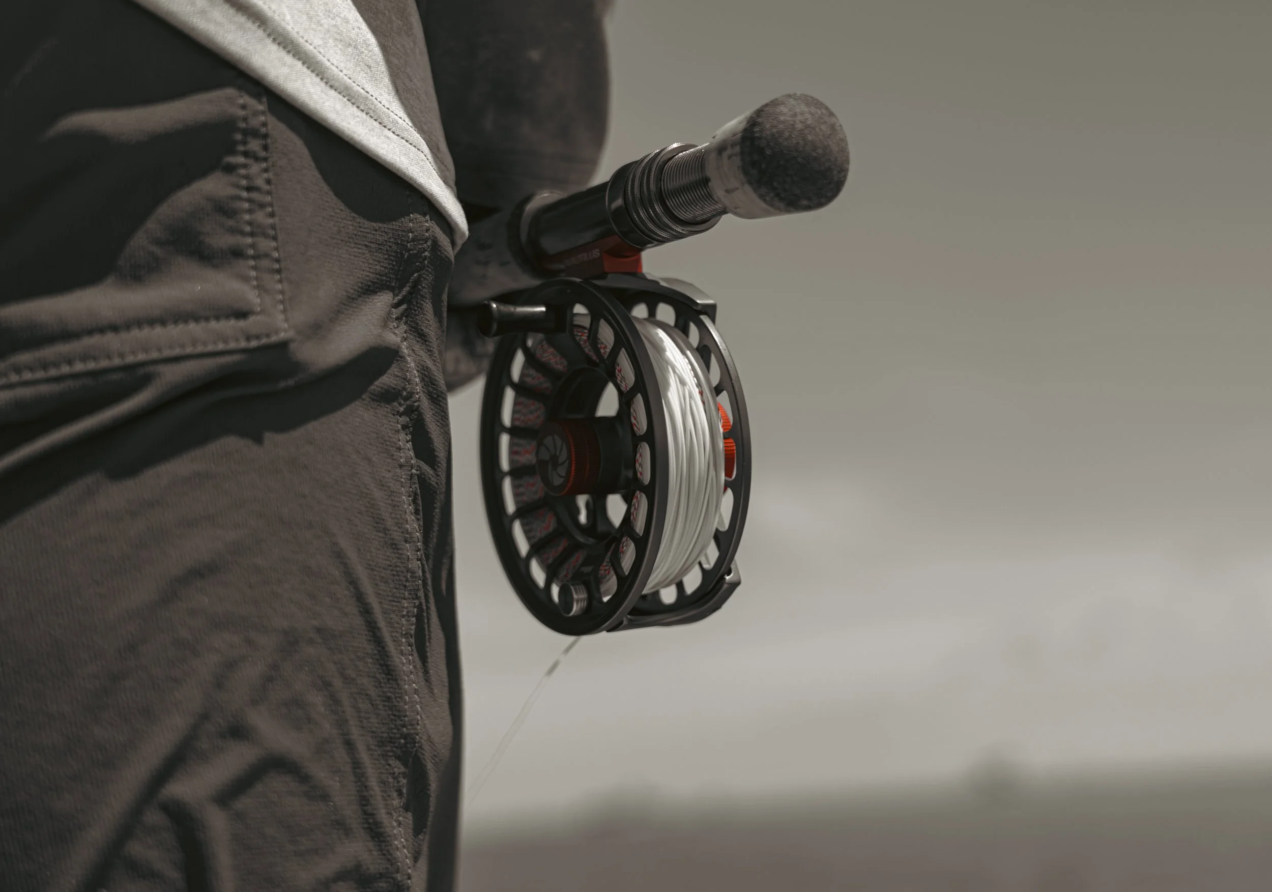 A person holding a fishing rod with a reel, wearing outdoor clothing, on a cloudy day.
