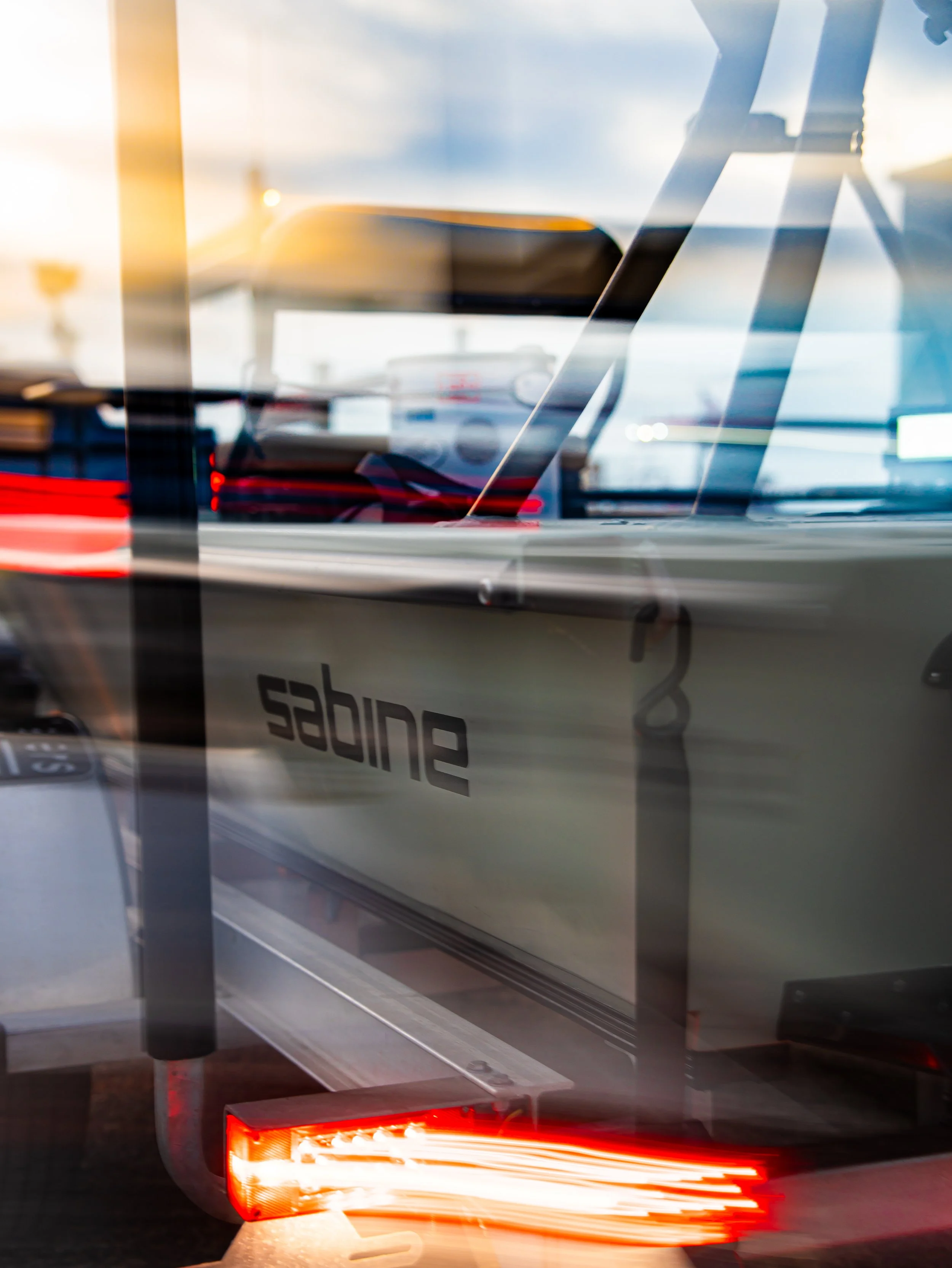 Close-up of a sailboat with the name 'Sabine' on its side, through a window with reflections of a sunset and possibly a car's taillights.
