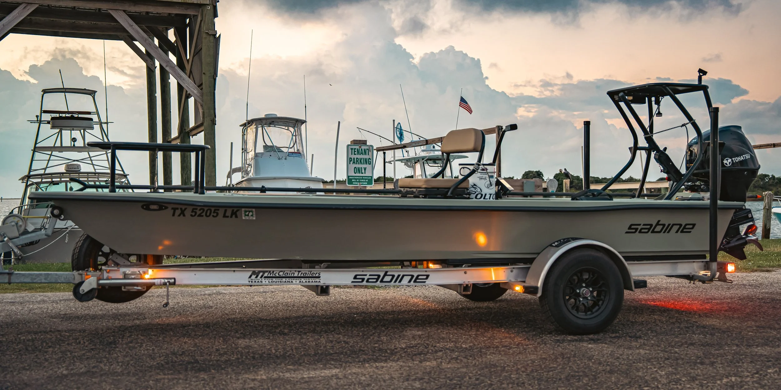 A boat on a trailer near a marina, with several boats docked in the background and a sunset sky overhead.