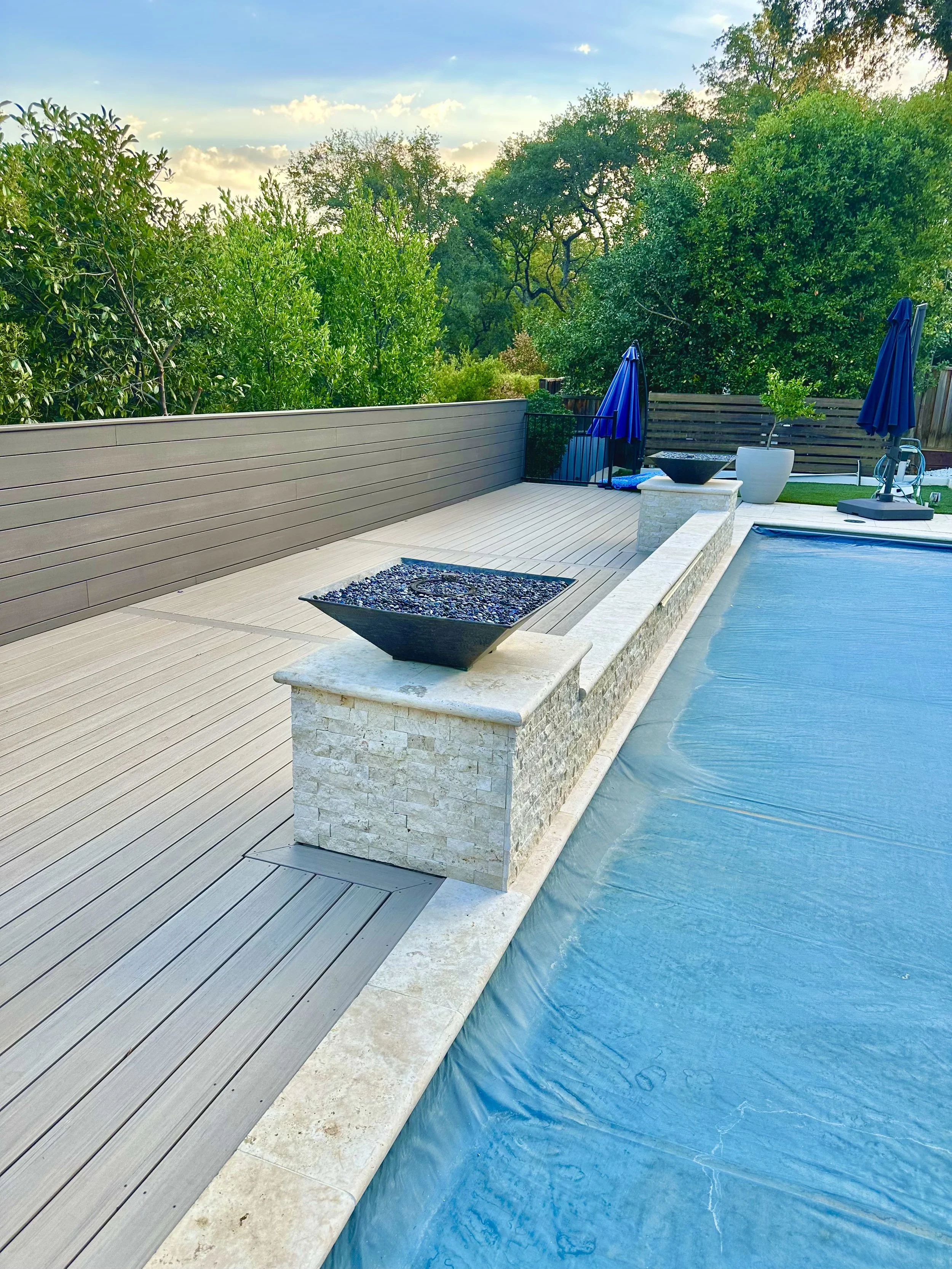 A backyard pool area with a blue pool cover, gray wooden deck, potted plants, and green trees in the background under a partly cloudy sky.