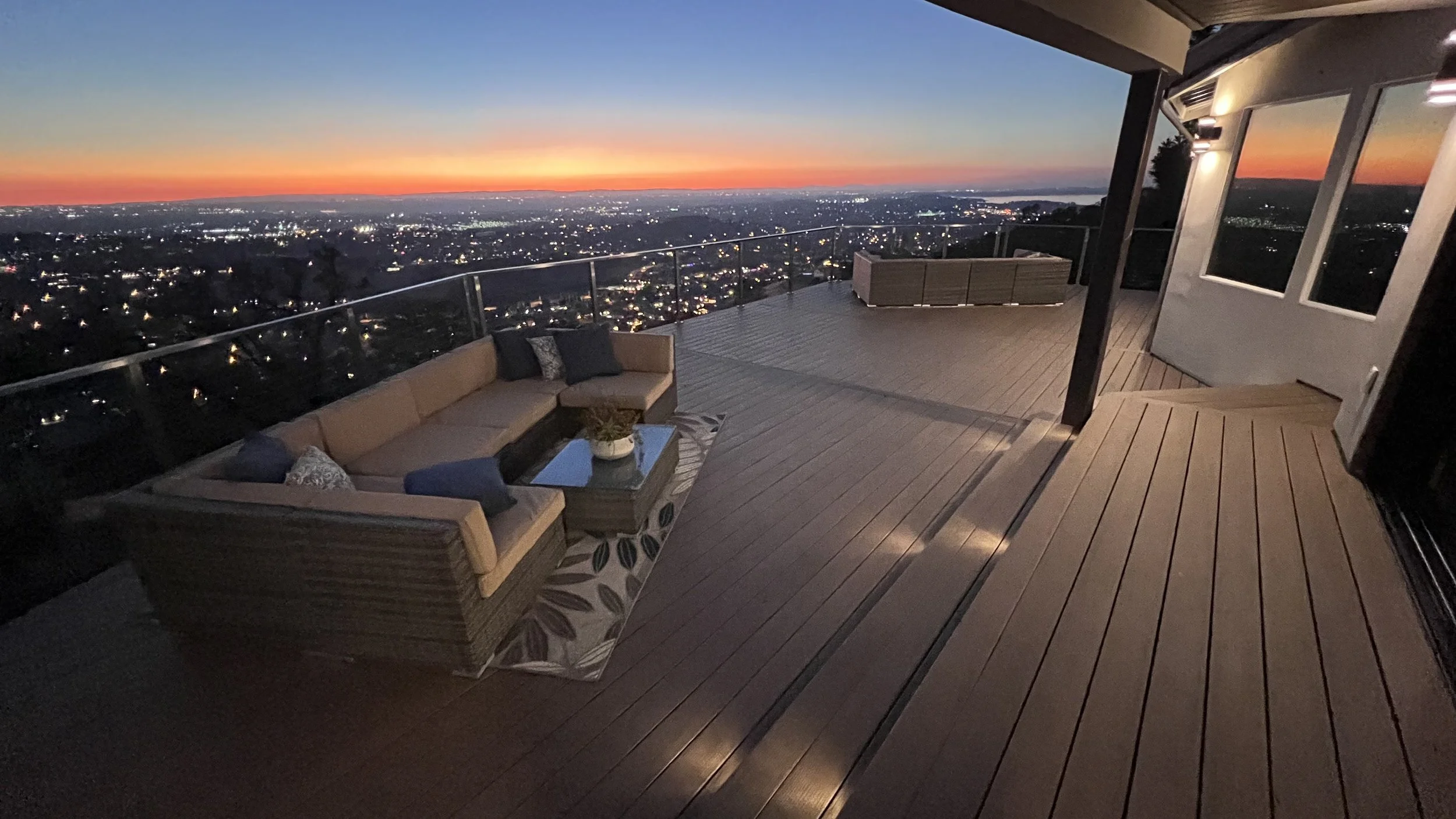 A rooftop deck at sunset with outdoor seating, a view of a cityscape, and a water body in the distance.