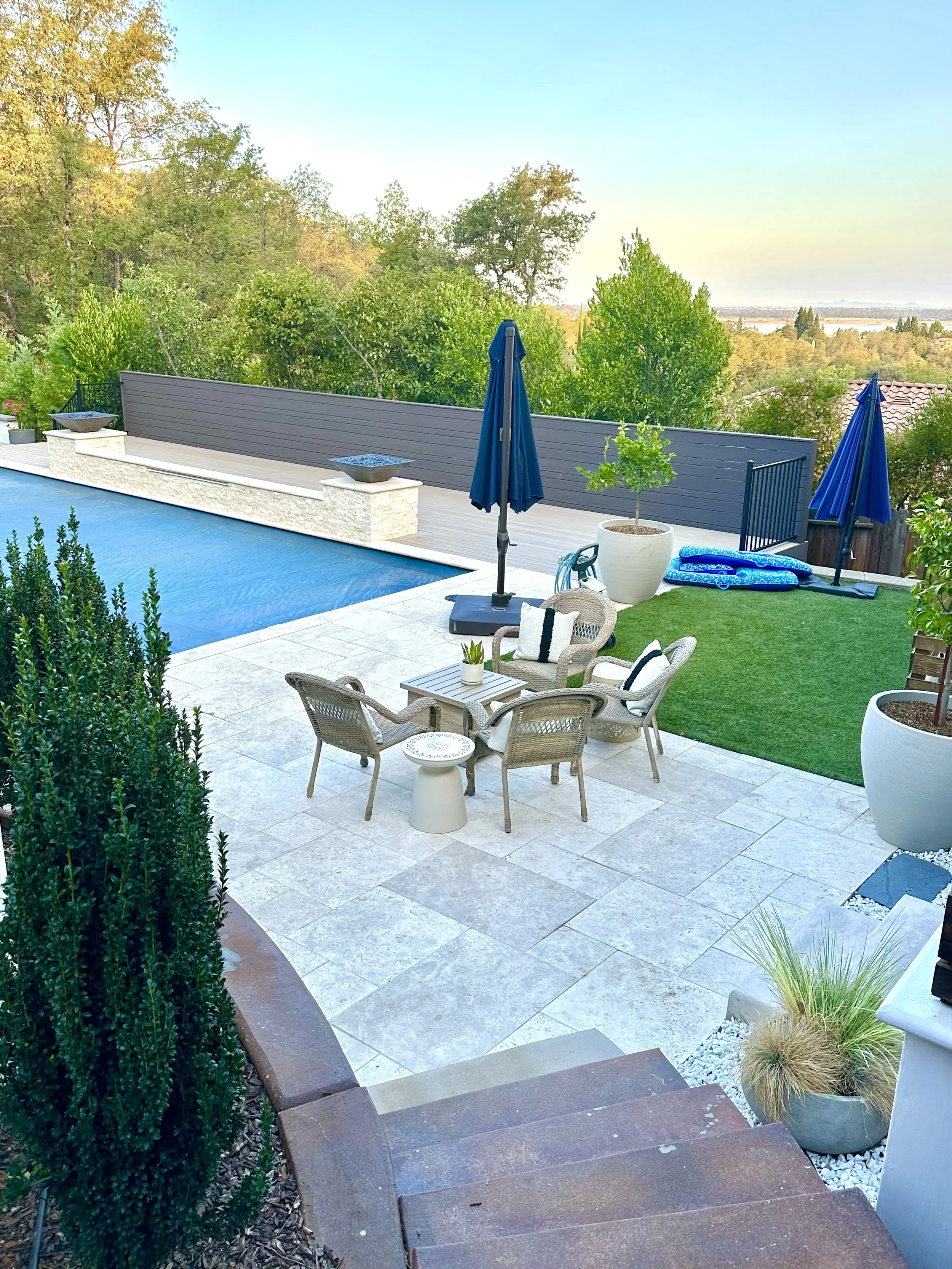 A backyard patio with a swimming pool, outdoor furniture, umbrellas, potted plants, and a view of trees and a distant landscape.
