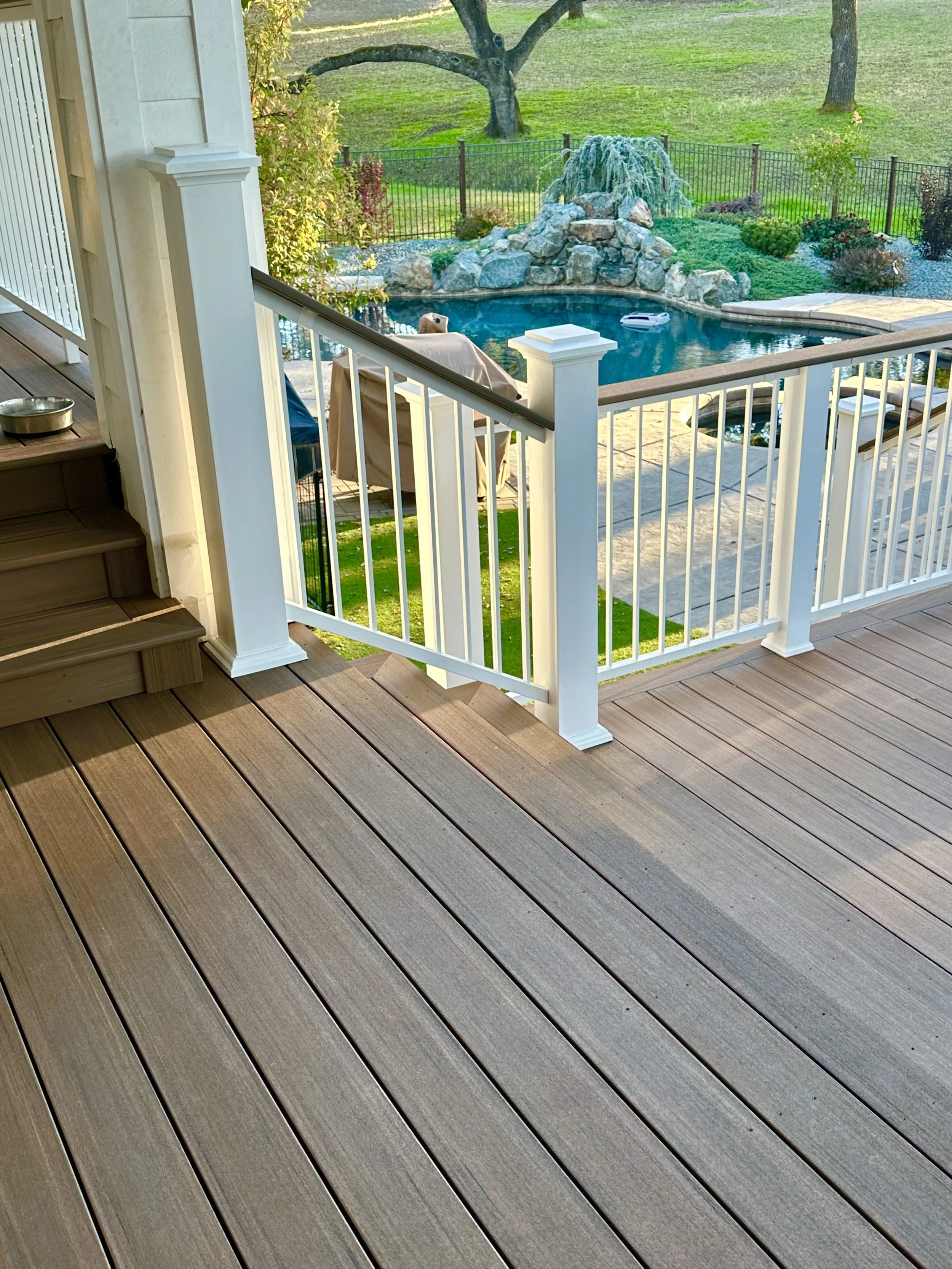 View from a wooden deck looking out over a backyard with a swimming pool, rocks, waterfall feature, outdoor furniture, trees, and a grassy lawn.