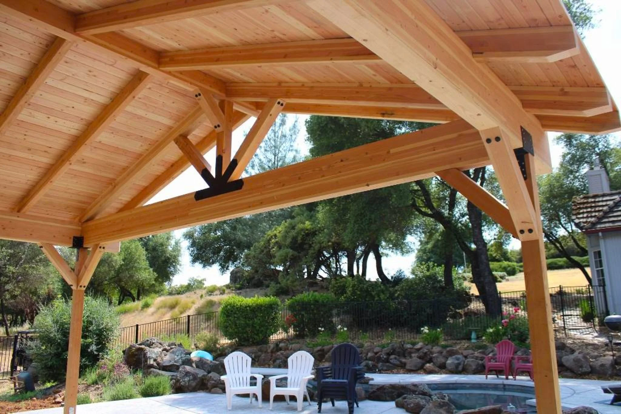 Under a wooden patio cover overlooking a backyard with a hot tub, white and black chairs, and a landscaped garden with trees and shrubs.