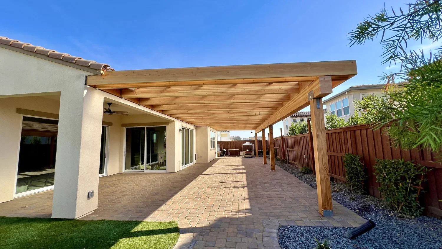 Backyard with a covered patio area, brick flooring, wooden pergola, sliding glass doors leading inside, wooden fence on the right, plants along the fence, and neighboring houses visible in the background.
