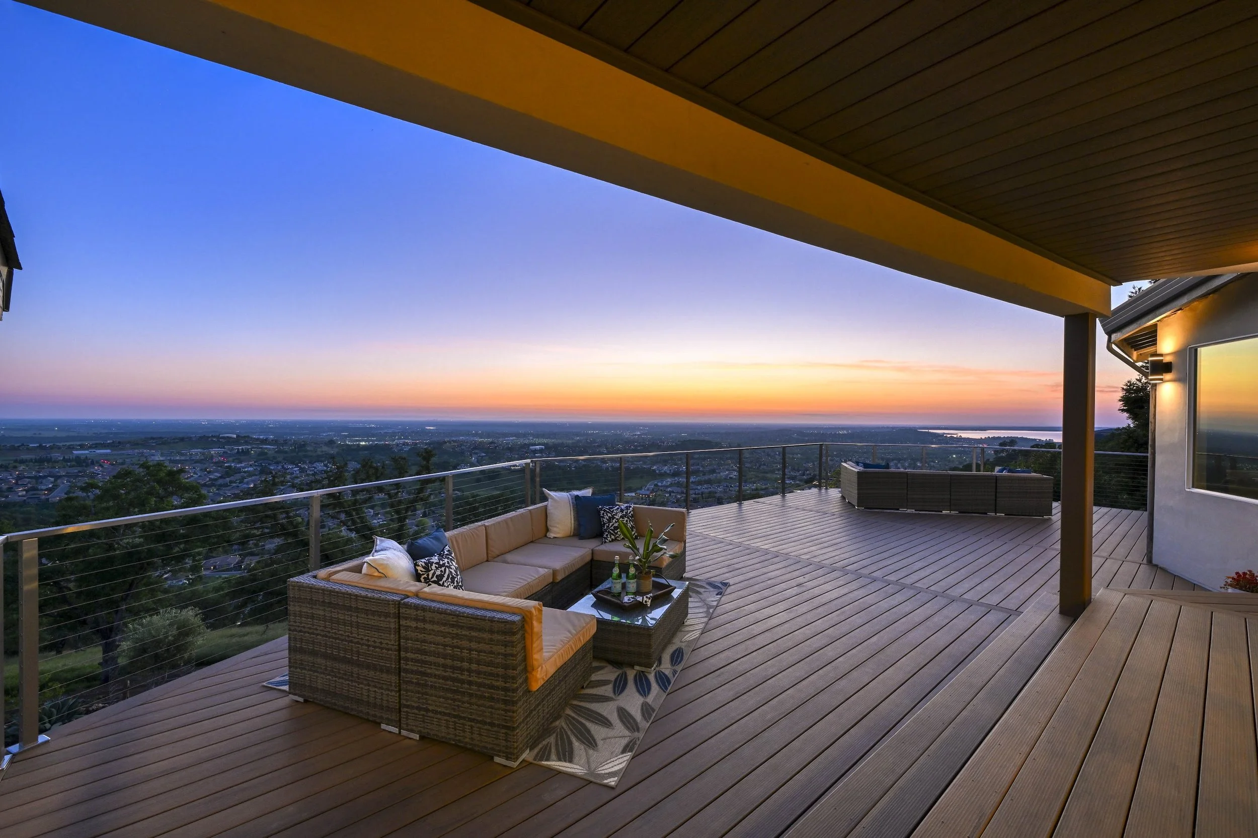 Empty wooden deck with a corner sofa and a coffee table, overlooking a scenic cityscape and sunset view.