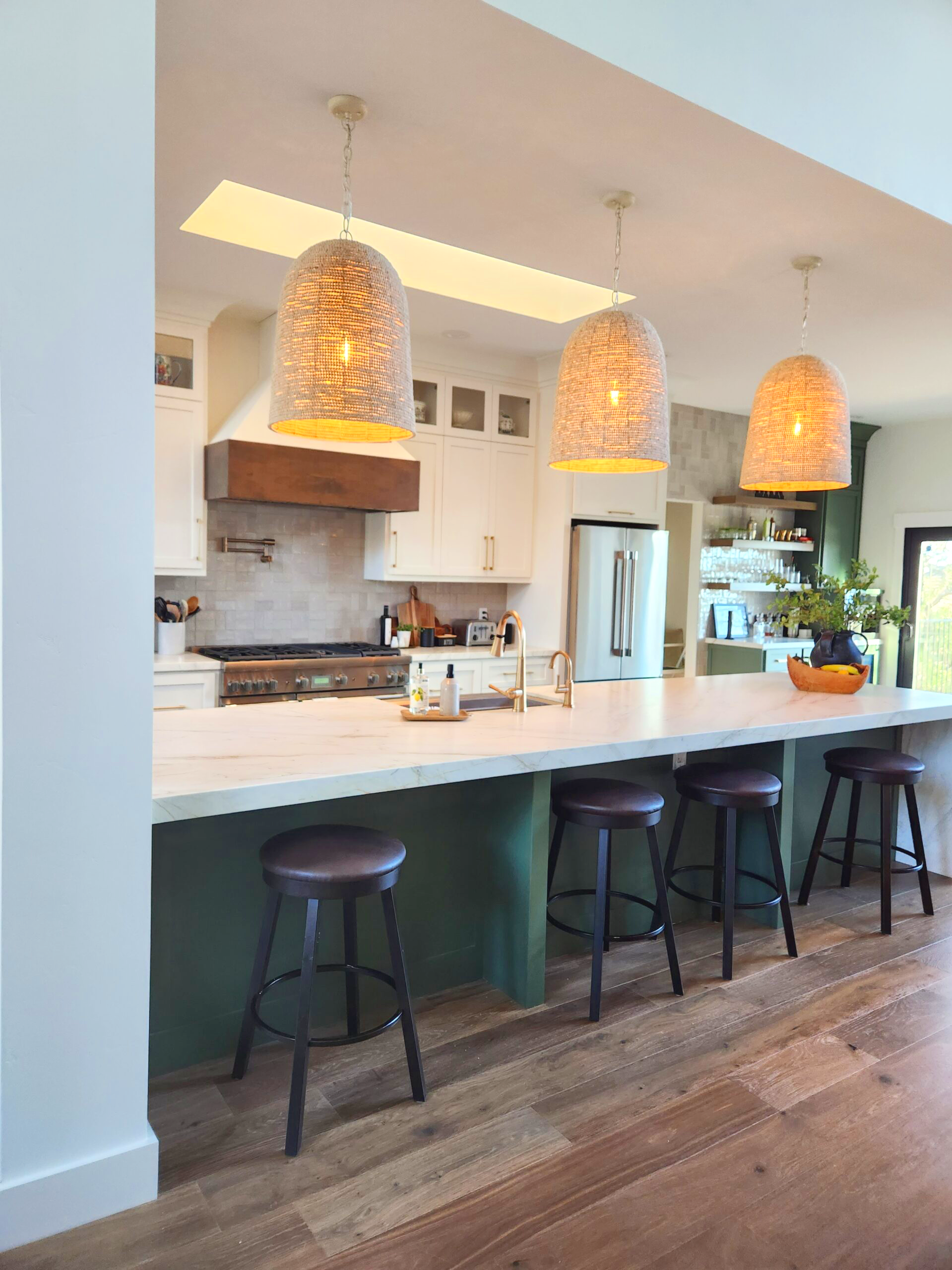 Kitchen with white cabinets, a large island with a white marble countertop, three hanging woven pendant lights, and black bar stools. There are stainless steel appliances, a wooden range hood, and greenery on the side.