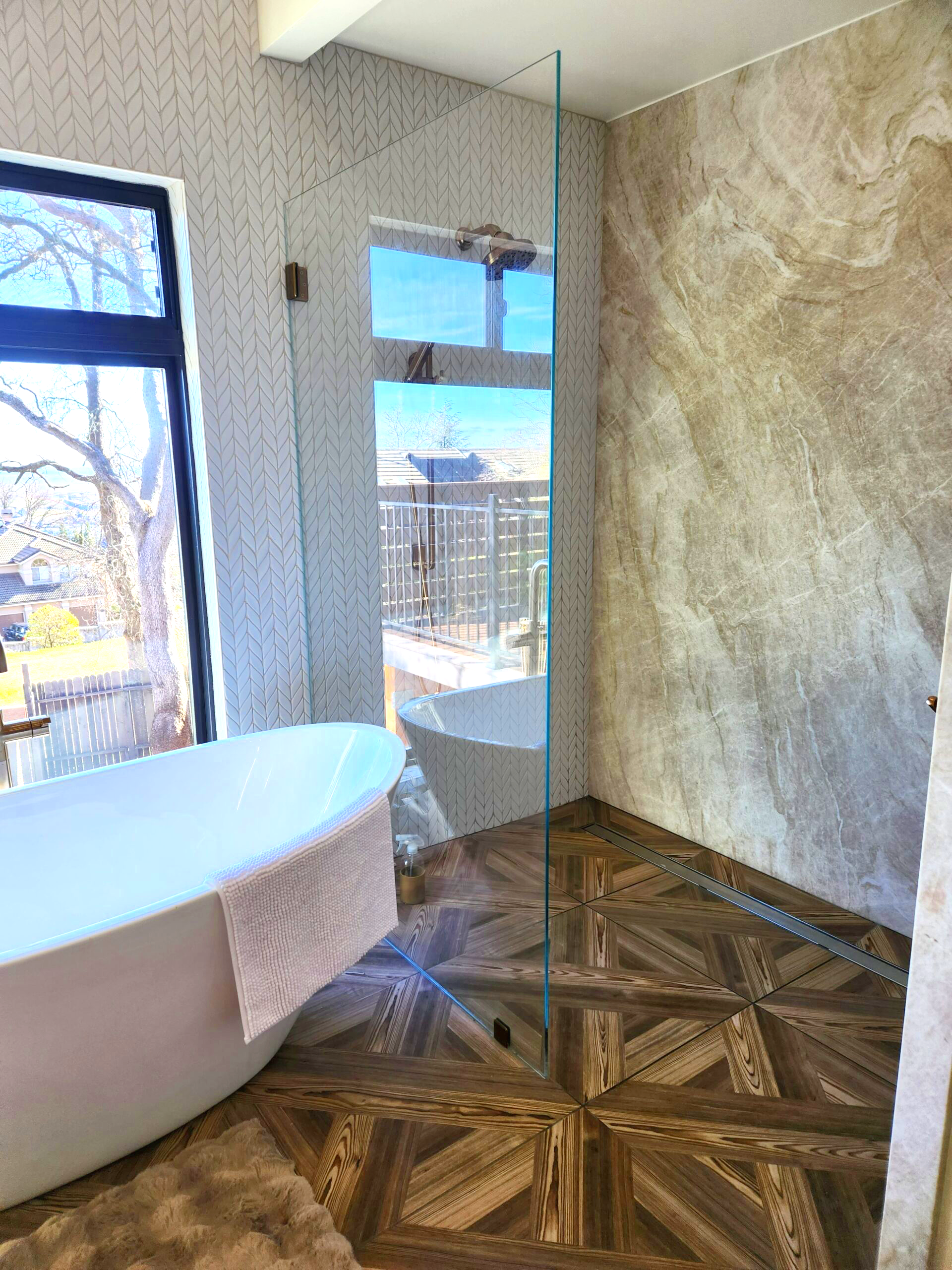 Bathroom with a freestanding bathtub, a large window, a glass shower partition, patterned wall tiles, and wood-patterned floor tiles.