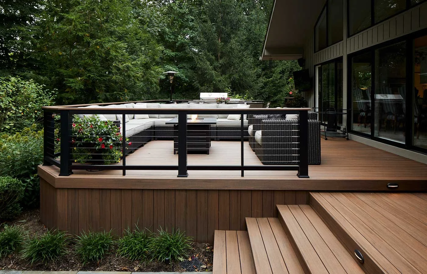 Wooden deck with black railing and outdoor seating, surrounded by greenery and trees.