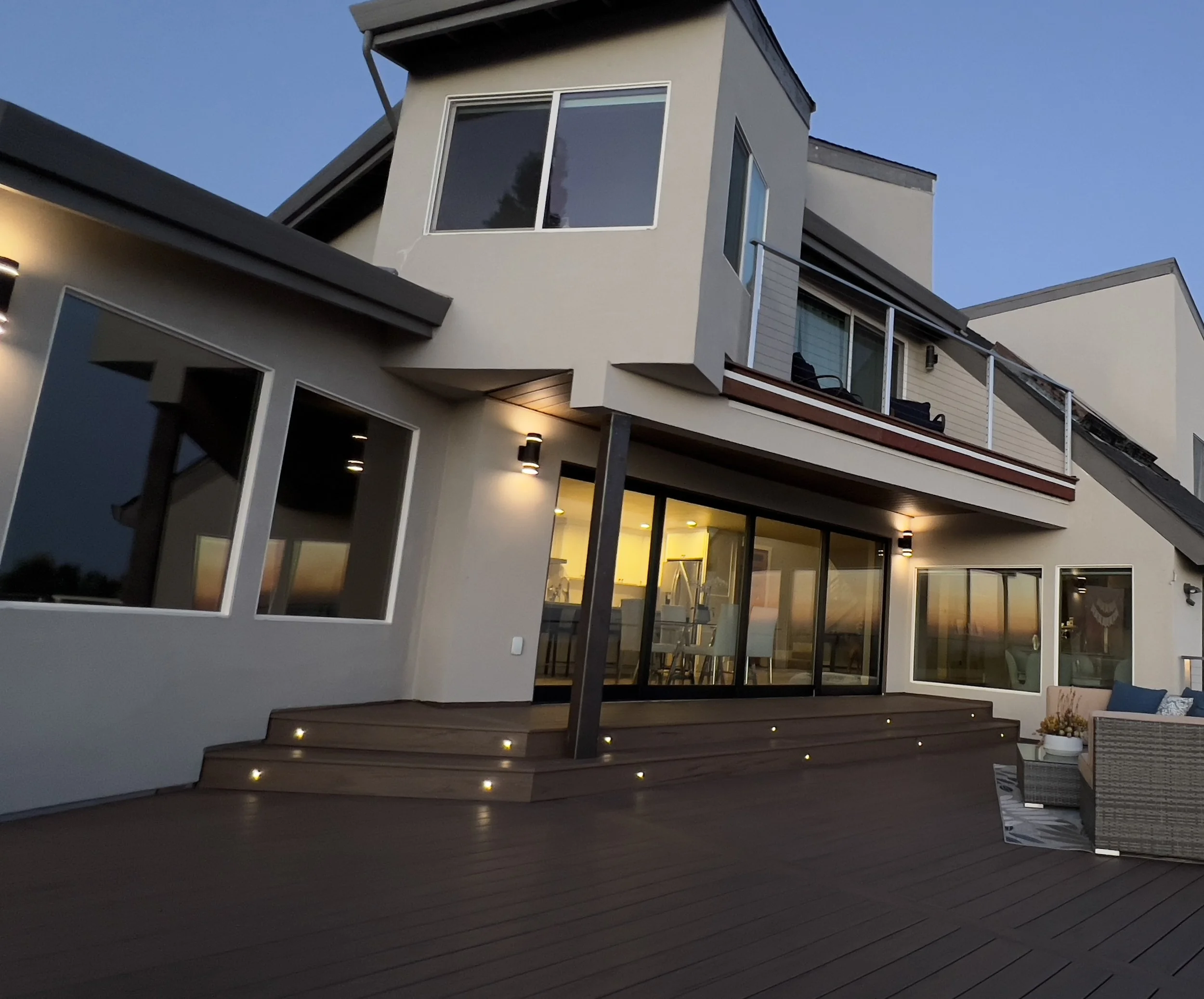Modern multi-story house with large windows and a spacious outdoor deck at dusk, illuminated by exterior lights.