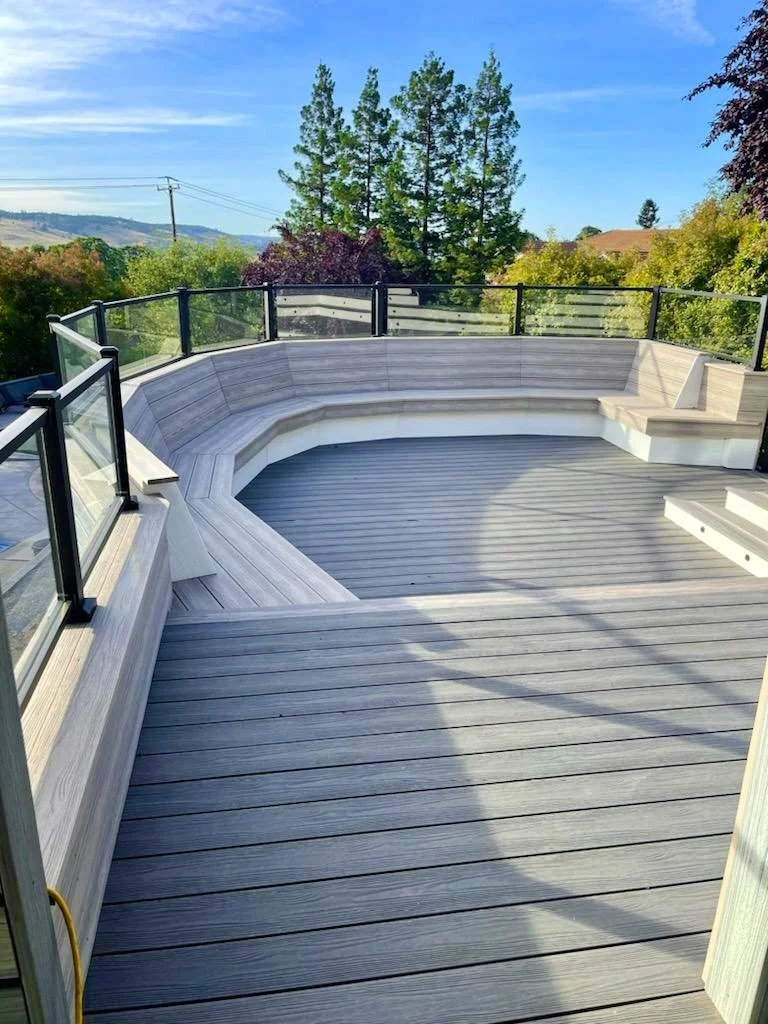 Modern outdoor deck with glass railing overlooking trees and hills under a blue sky.