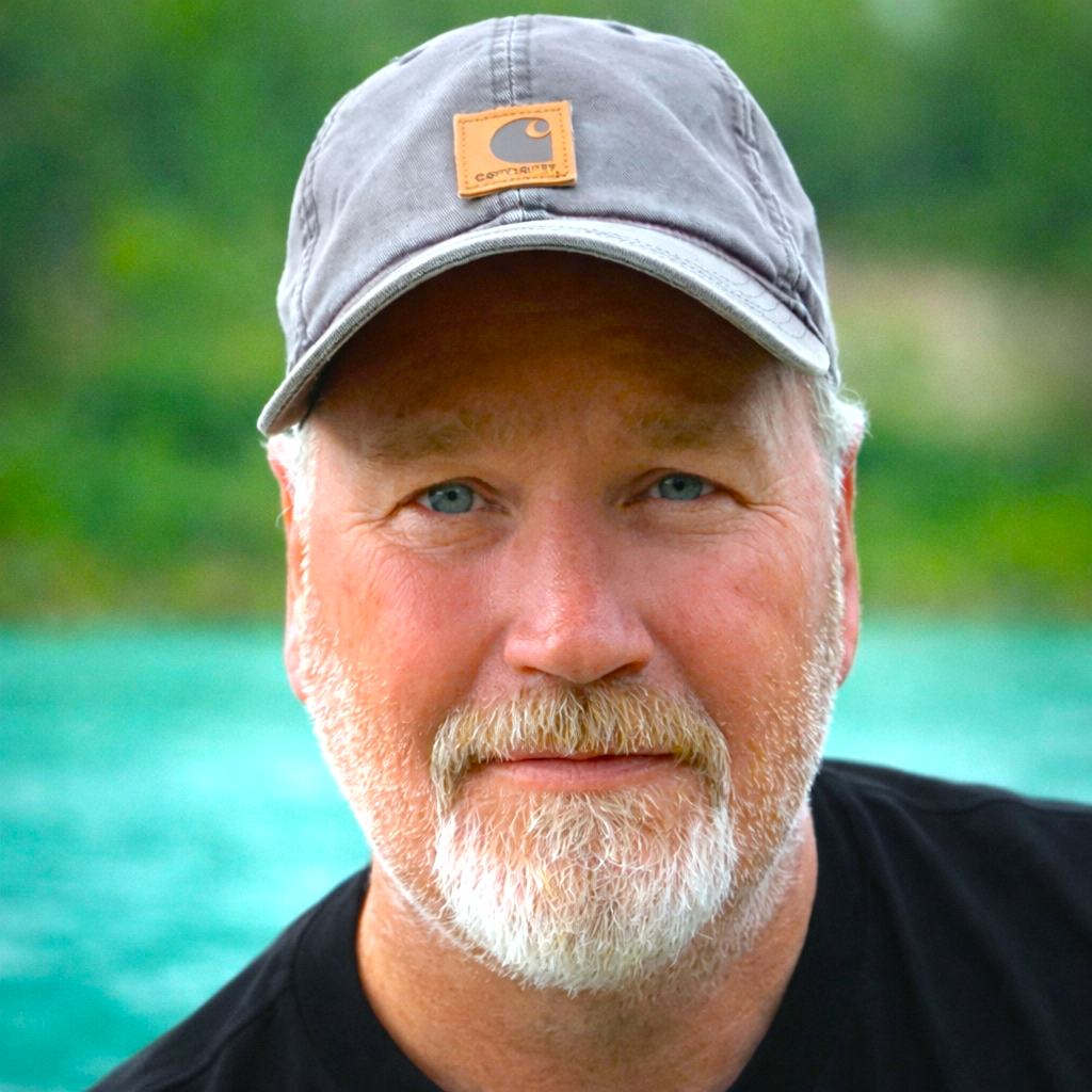 Close-up of a man with a beard and mustache wearing a gray Carhartt cap, outdoors near a body of water with green trees and cliffs in the background.