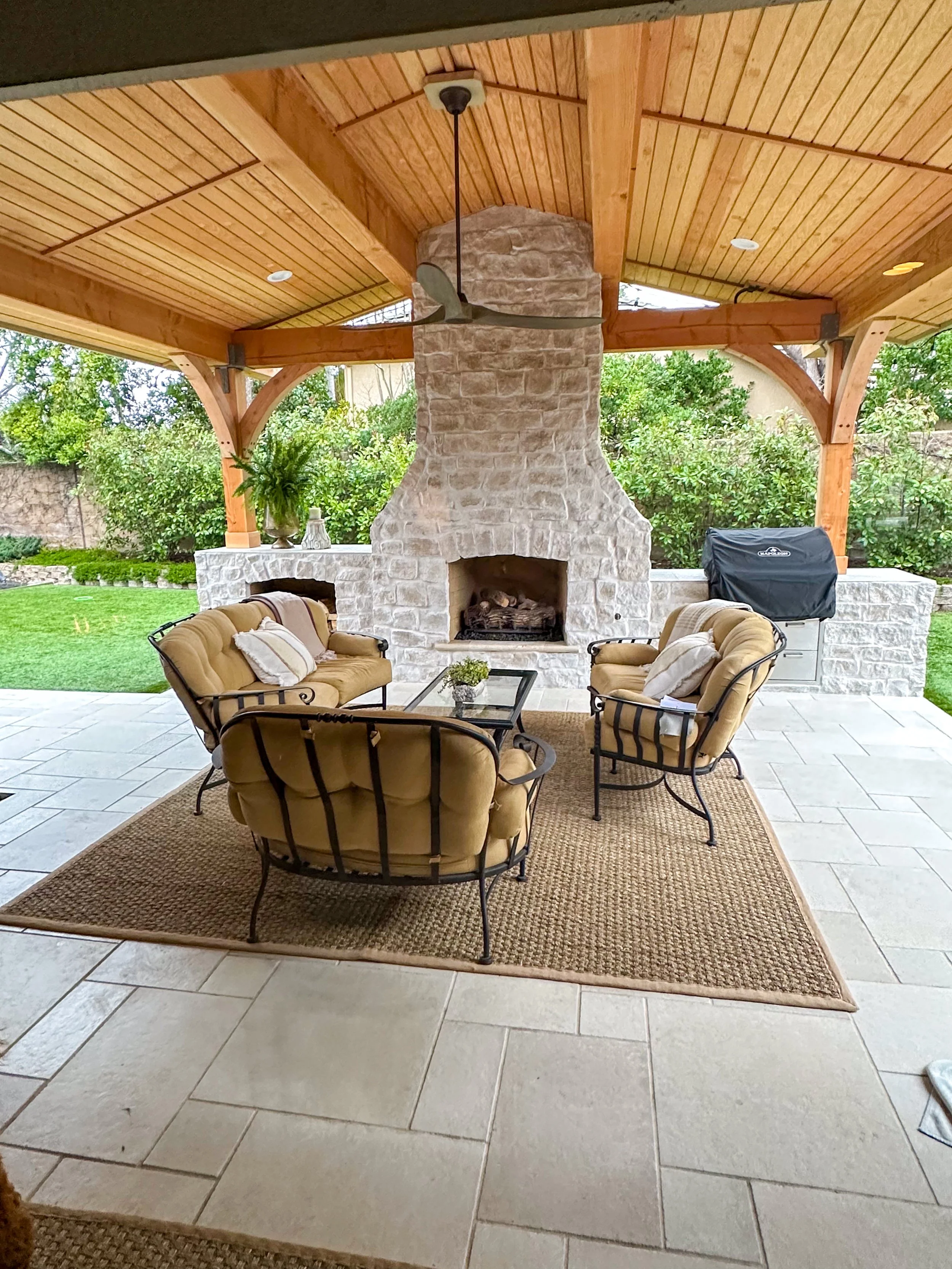 Outdoor living space with a stone fireplace, cushioned patio furniture, a roof with wood paneling, ceiling fan, and greenery in the background.