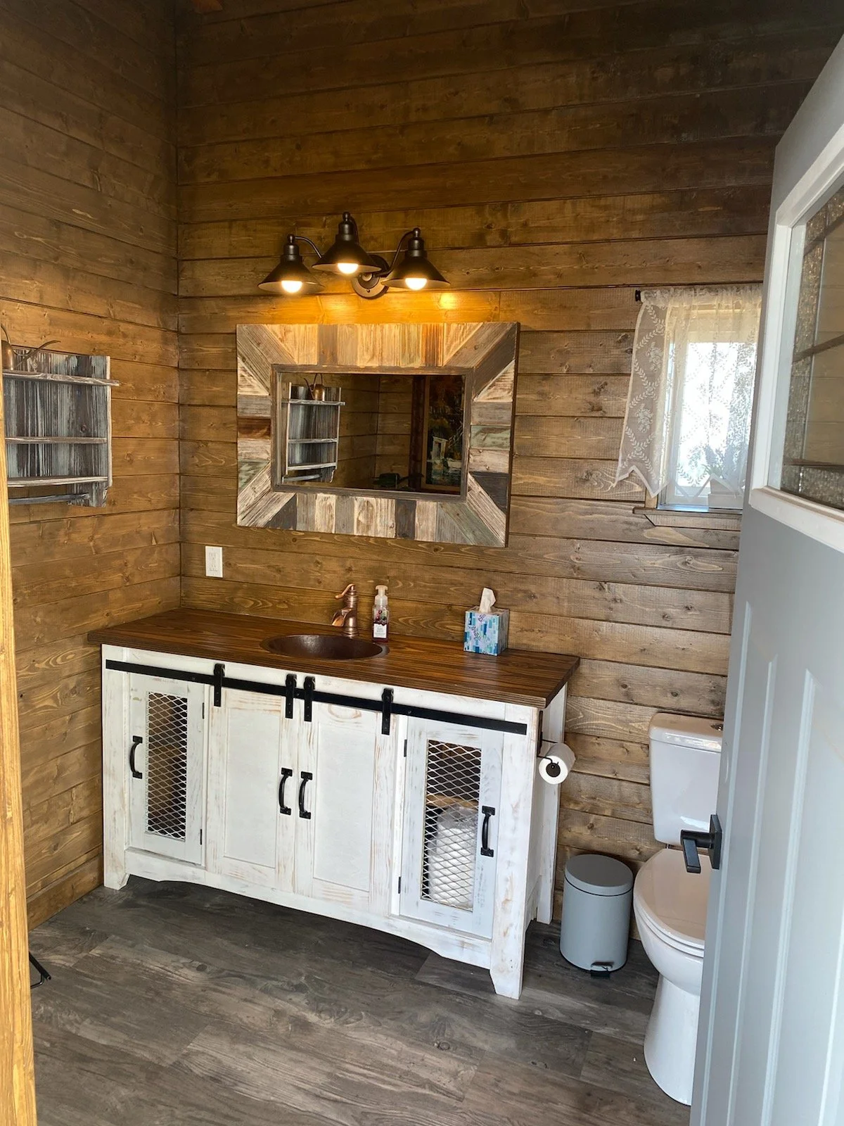 Rustic bathroom with wood-paneled walls, a white distressed vanity with black hardware, a small copper sink, a wooden countertop, a large decorative mirror, a wall-mounted light fixture, a small window with lace curtain, and a toilet with a trash can
