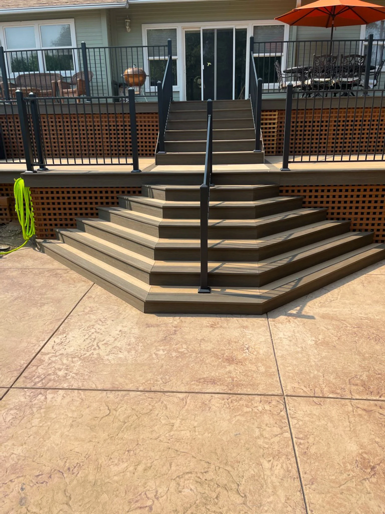 Wooden deck stairs with black metal railings leading up to an elevated balcony. The deck has outdoor furniture, an orange umbrella, and a grill, with a house in the background.