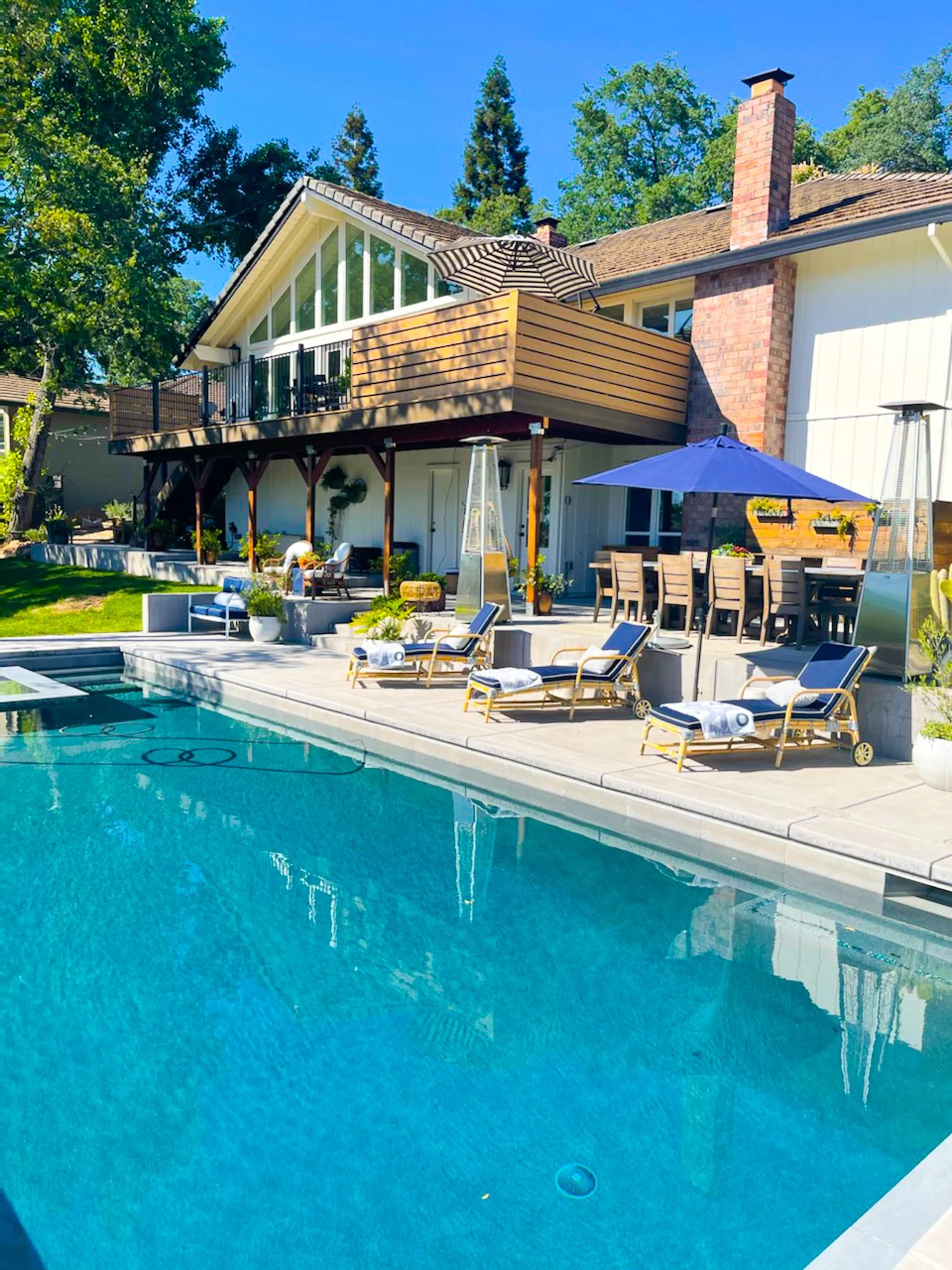 Luxury backyard with swimming pool, lounge chairs, patio dining area, and a house with a deck, large windows, and a brick chimney, surrounded by trees.