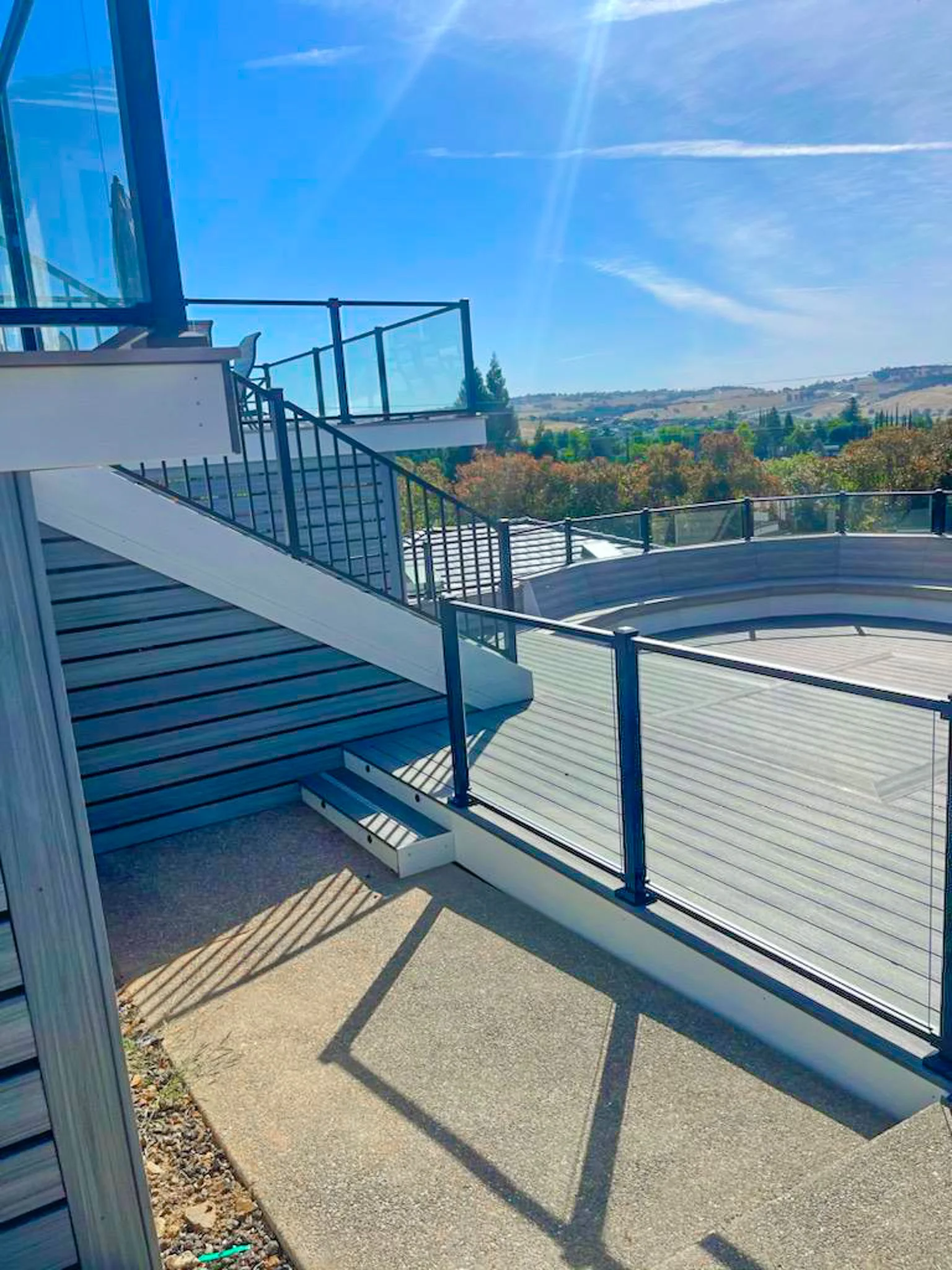 View of a balcony with metal railings, stairs, and a circular hot tub, overlooking a landscape with trees and hills under a blue sky.